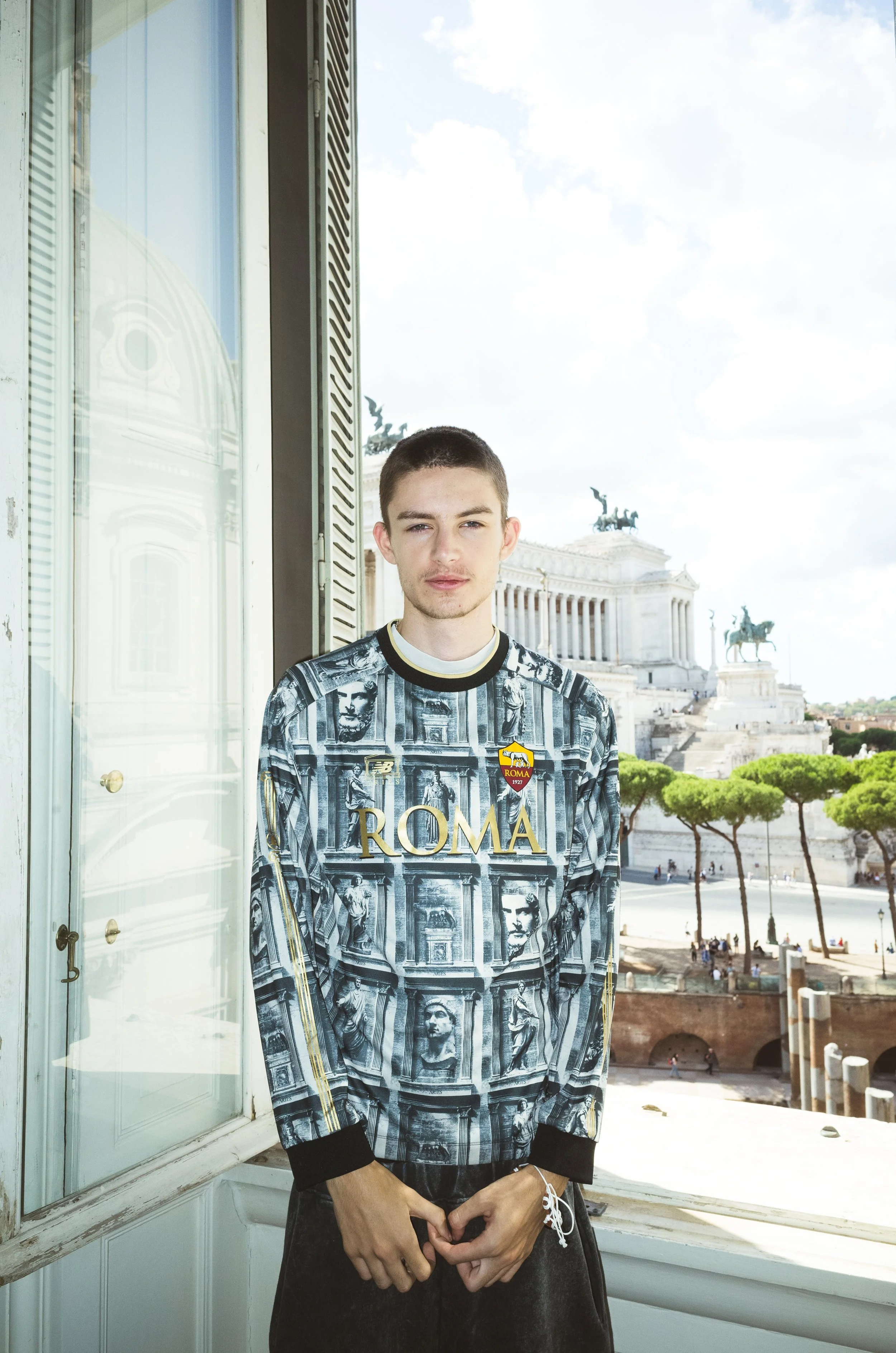 A young man standing near a window with the Altare della Patria and statues in Rome in the background.