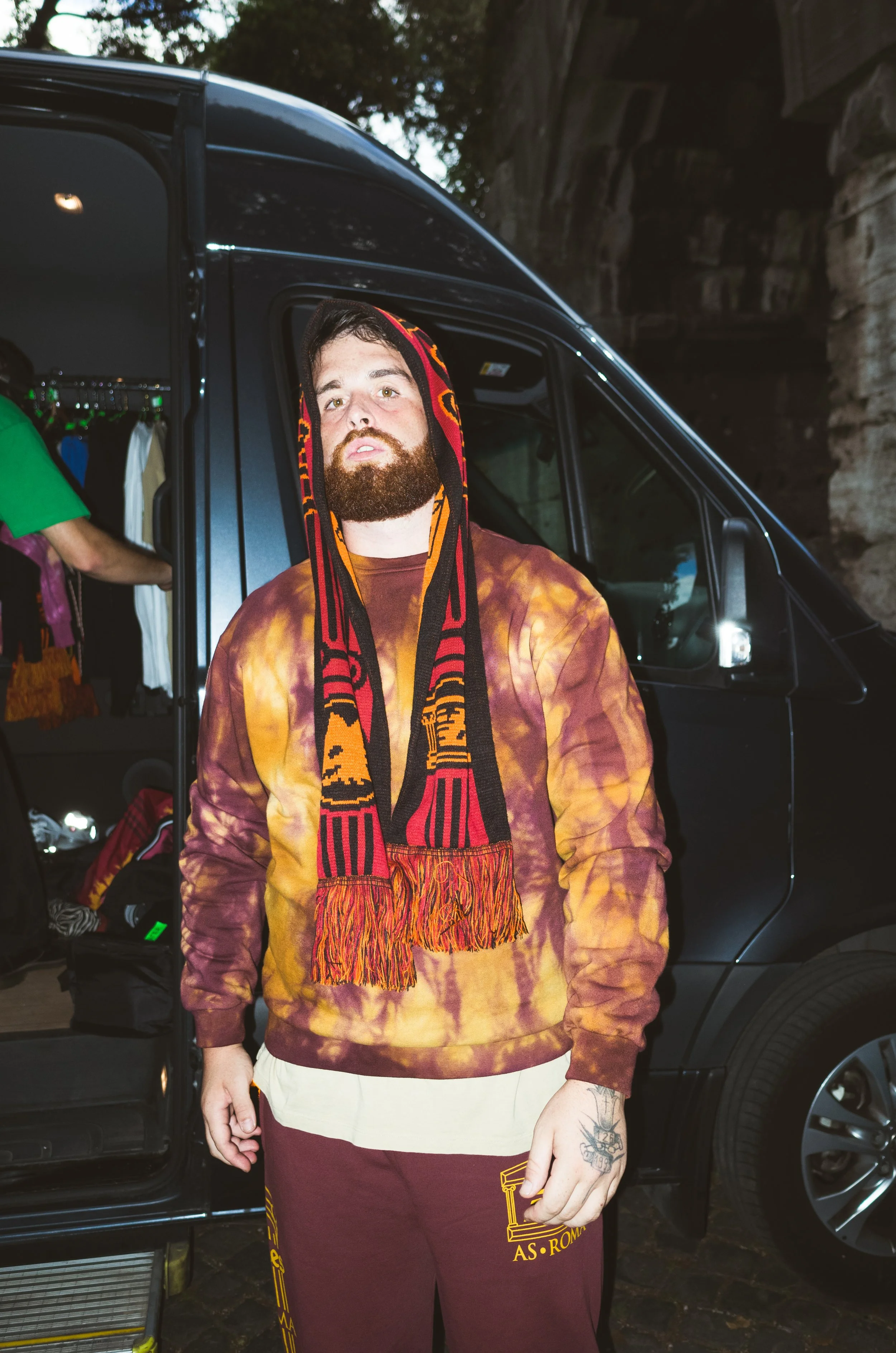 A man with a beard wearing a tie-dye hoodie and a red, orange, and black scarf stands outside near a black van with an open door, with a person in an orange shirt visible inside the van.