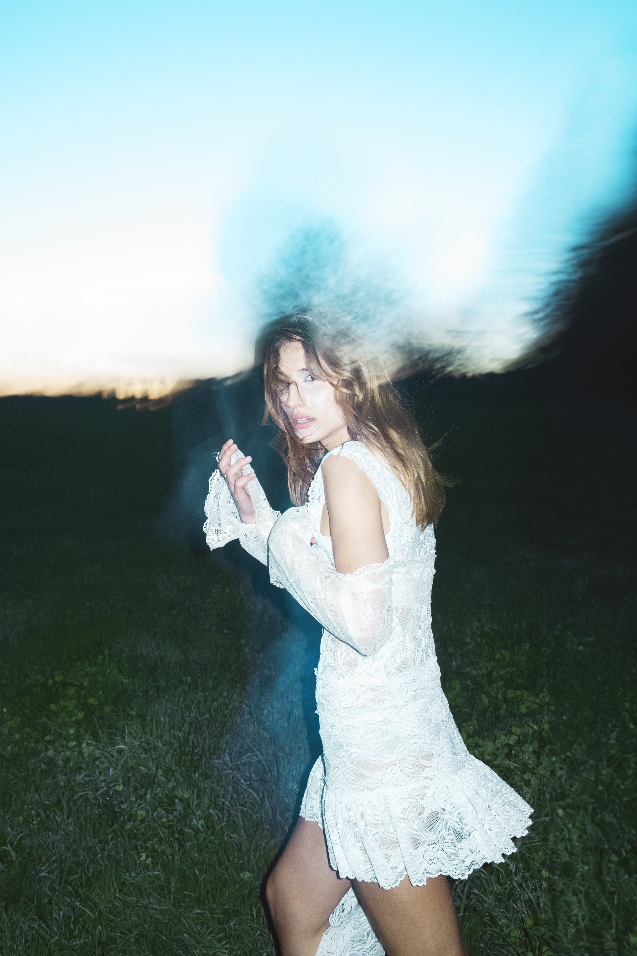 Young woman in a white lace dress standing outdoors during twilight, with motion blur around her, creating a dynamic and artistic effect.