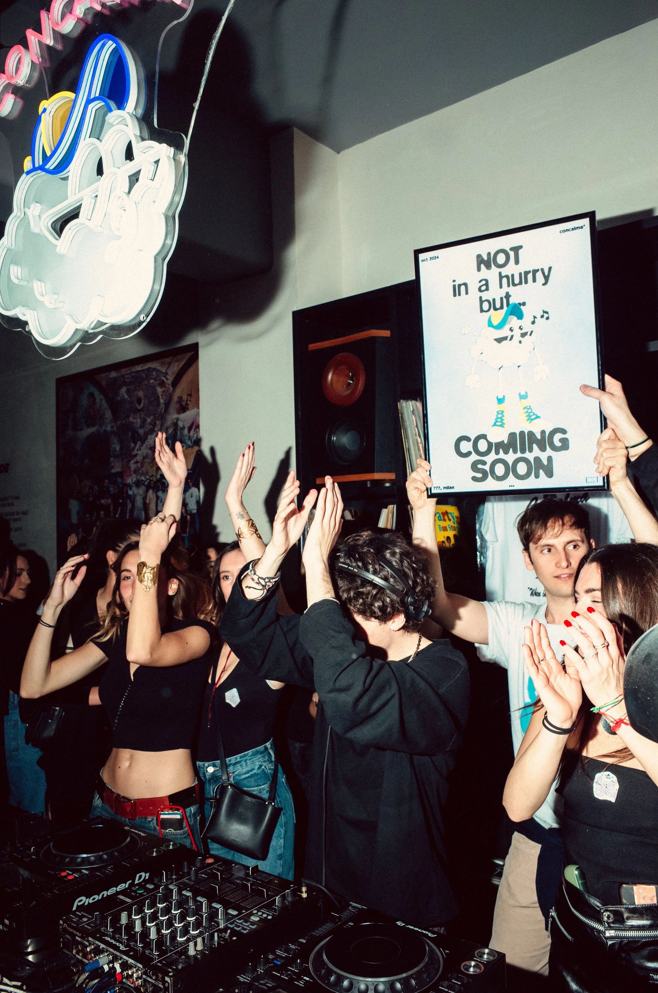 Group of people dancing at a party or club, with a DJ in the foreground and a sign that reads 'Not in a hurry but coming soon' in the background.