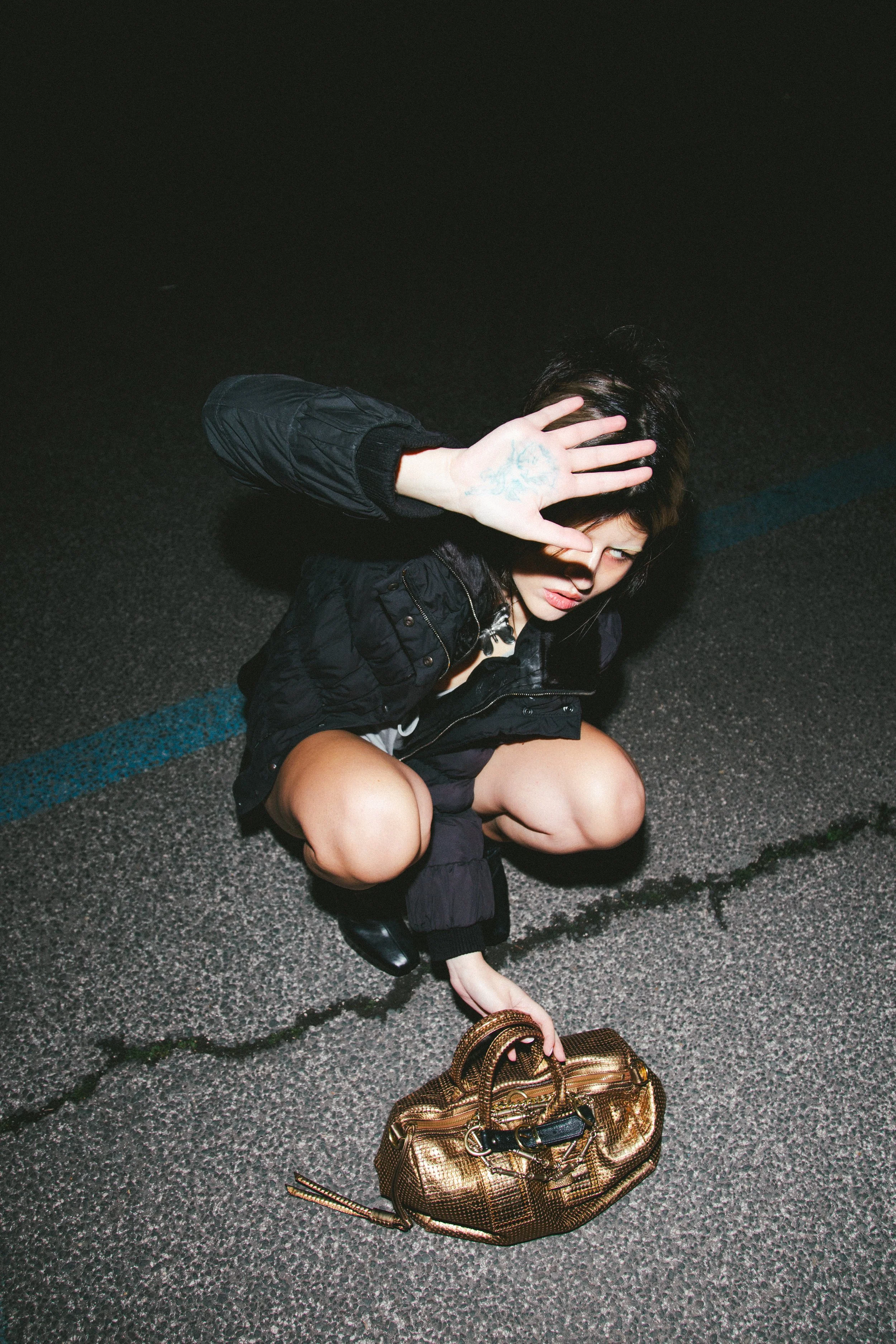 A woman squatting on the street at night, covering her eyes with one hand, holding a shiny gold purse with the other.
