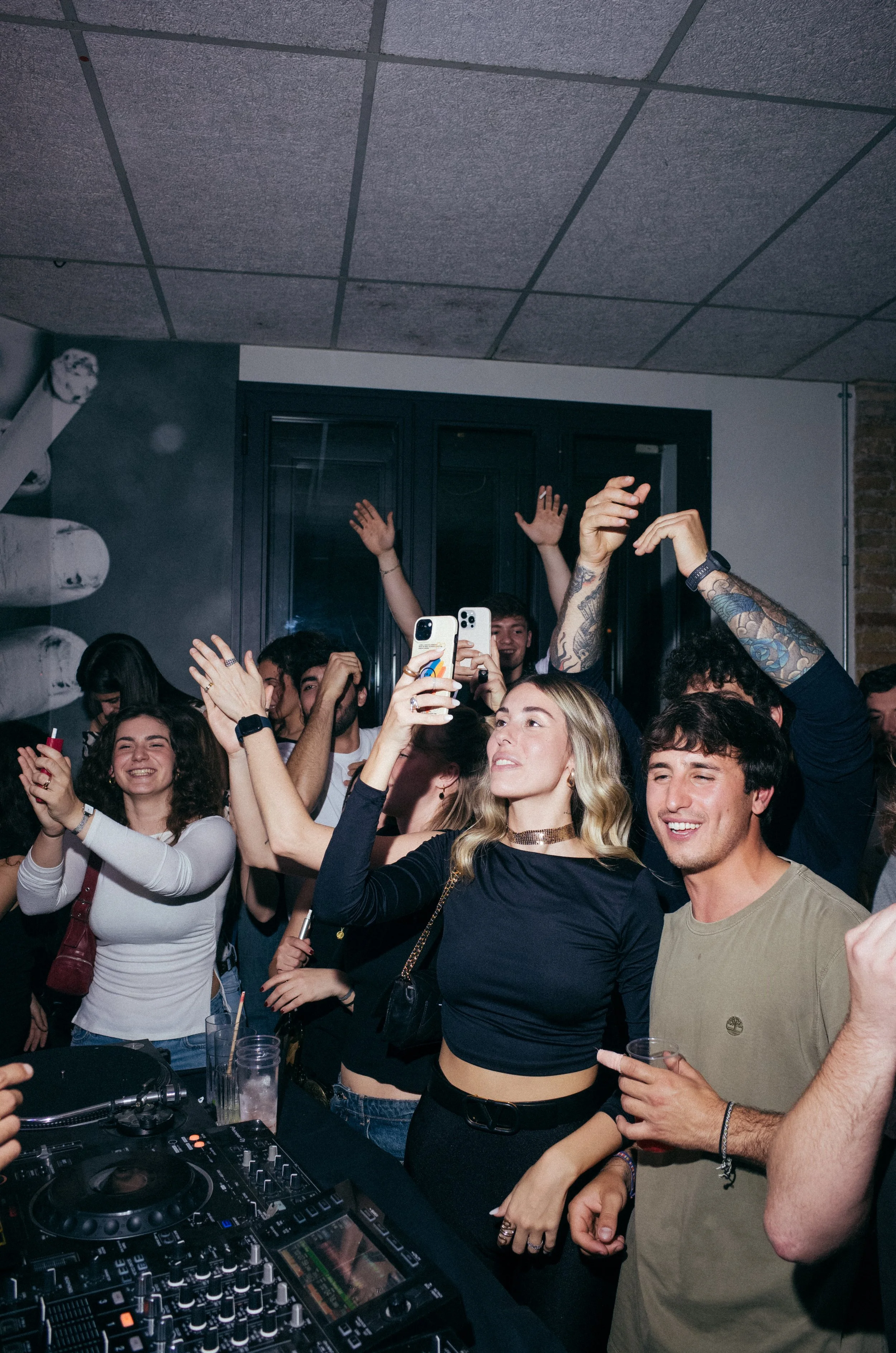 Group of young people enjoying a party, some with drinks, dancing, and taking pictures near DJ equipment.
