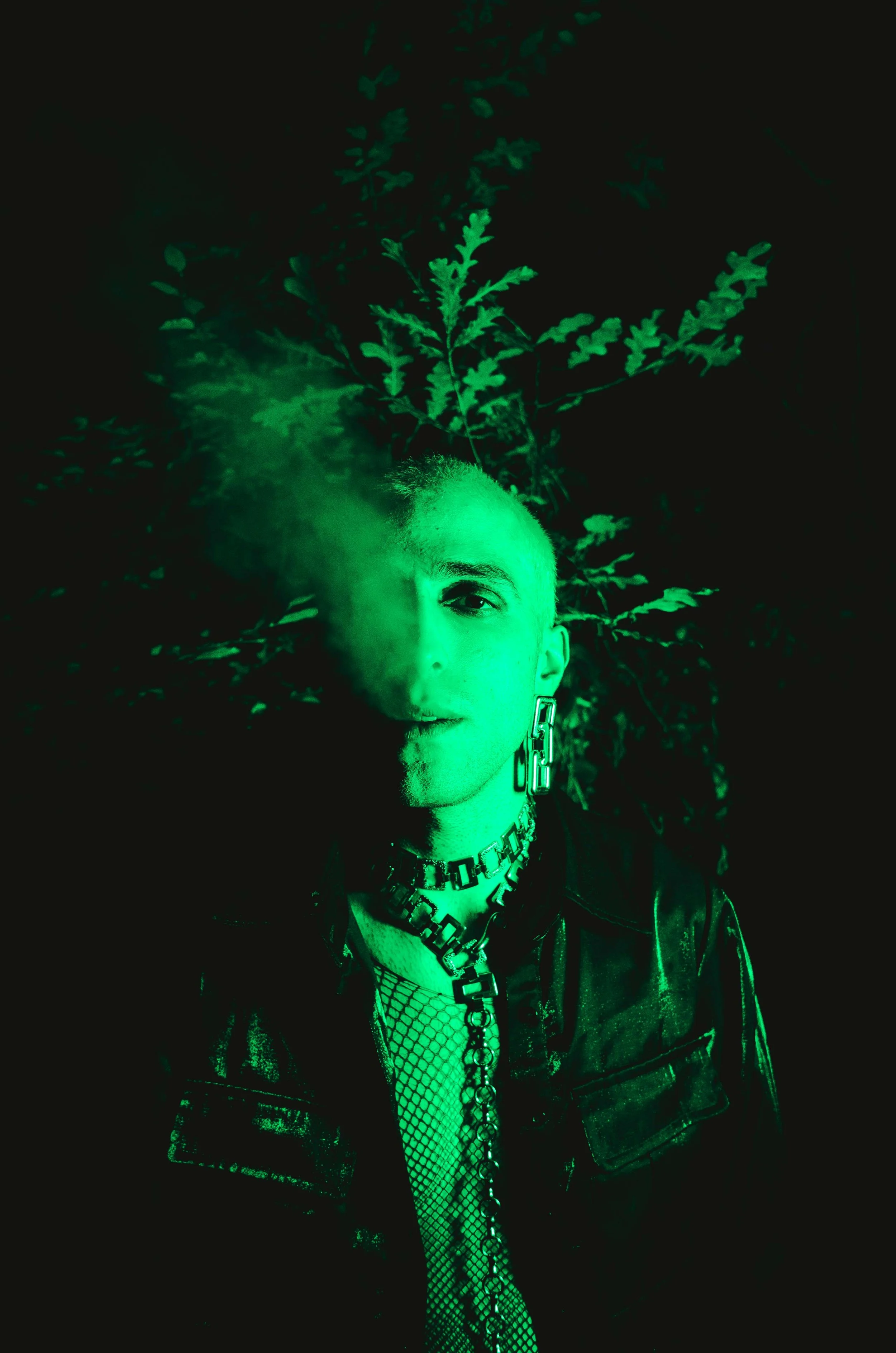Person with short hair, dressed in black leather jacket, mesh top, and chunky jewelry, standing in front of a leafy background, with green neon lighting and smoke effect.