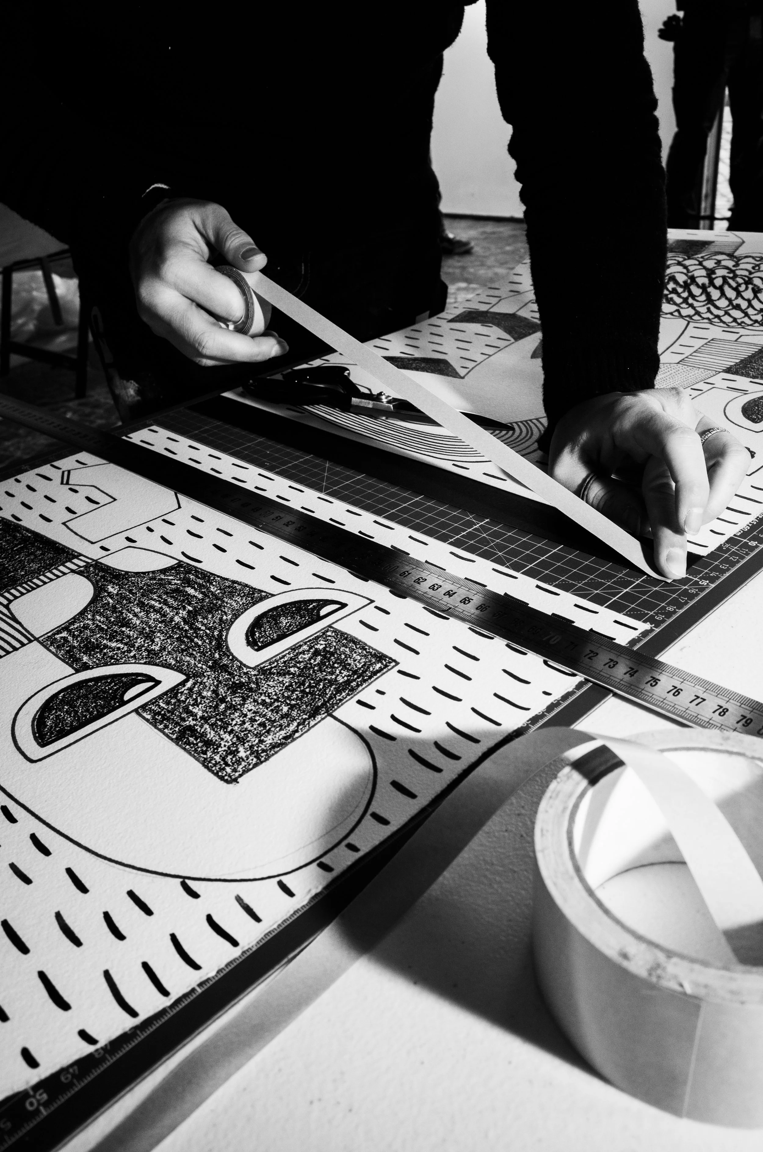 Person measuring and tracing a graphic design on paper with a ruler and masking tape on a desk.
