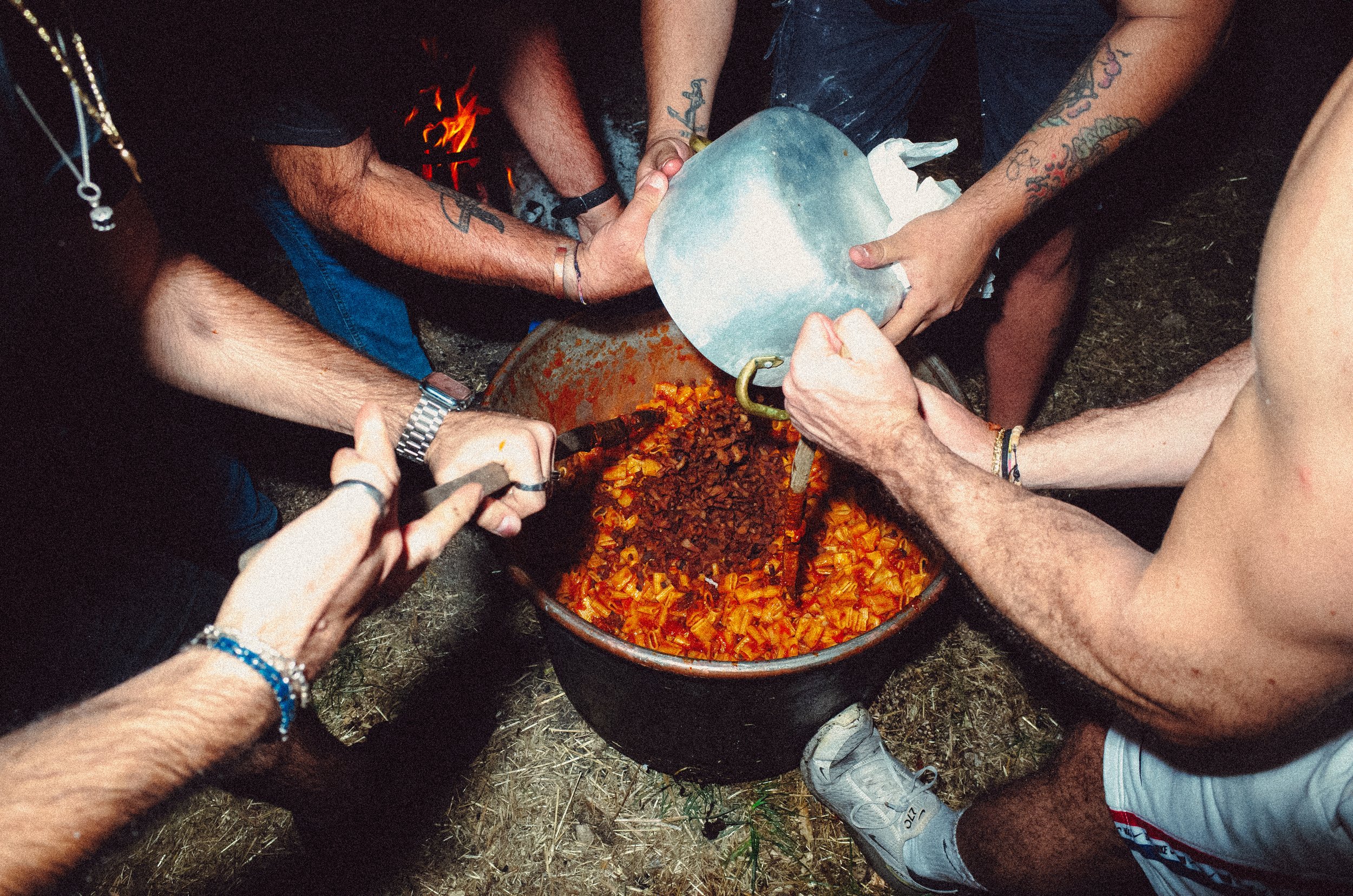 Group of people preparing a large pot of chili outdoors at night, stirring the mixture together.