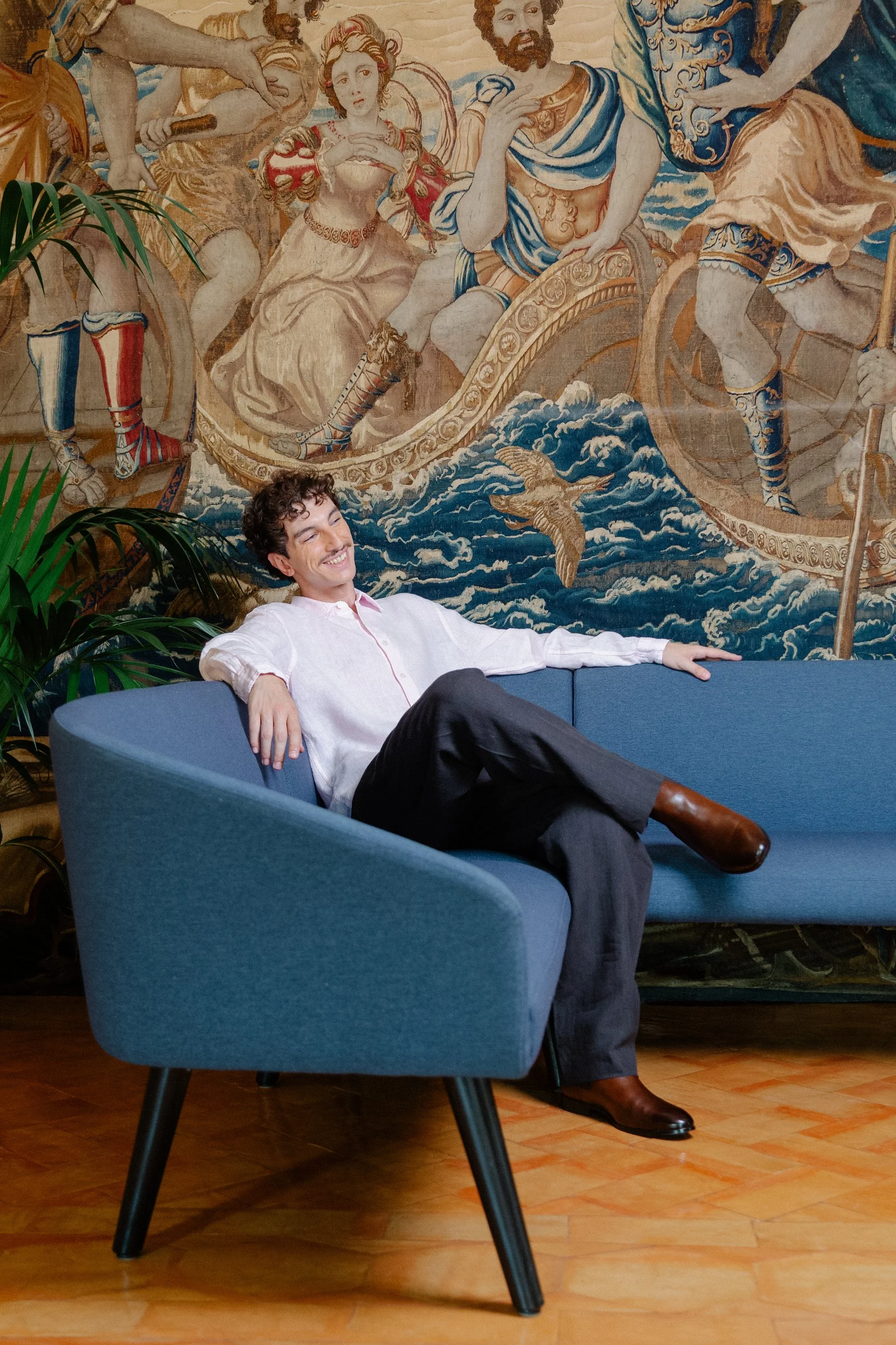 A man with curly hair, wearing a white button-down shirt and dark pants, sitting on a blue sofa with a smile, in front of a large ancient mural depicting mythological figures riding chariots over the sea.