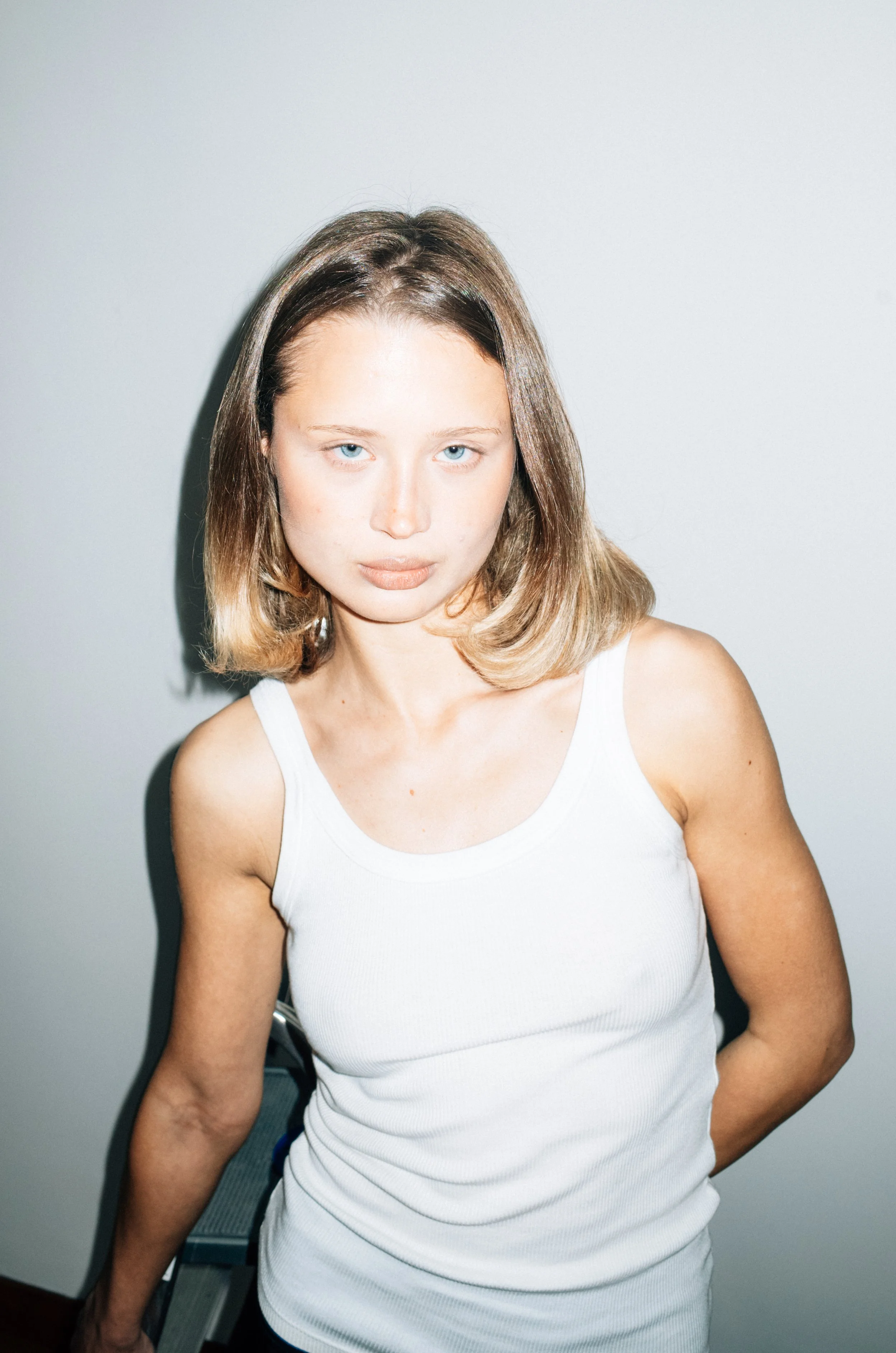 Portrait of a young woman with shoulder-length light brown hair and blue eyes, wearing a white sleeveless top, standing against a plain white wall.