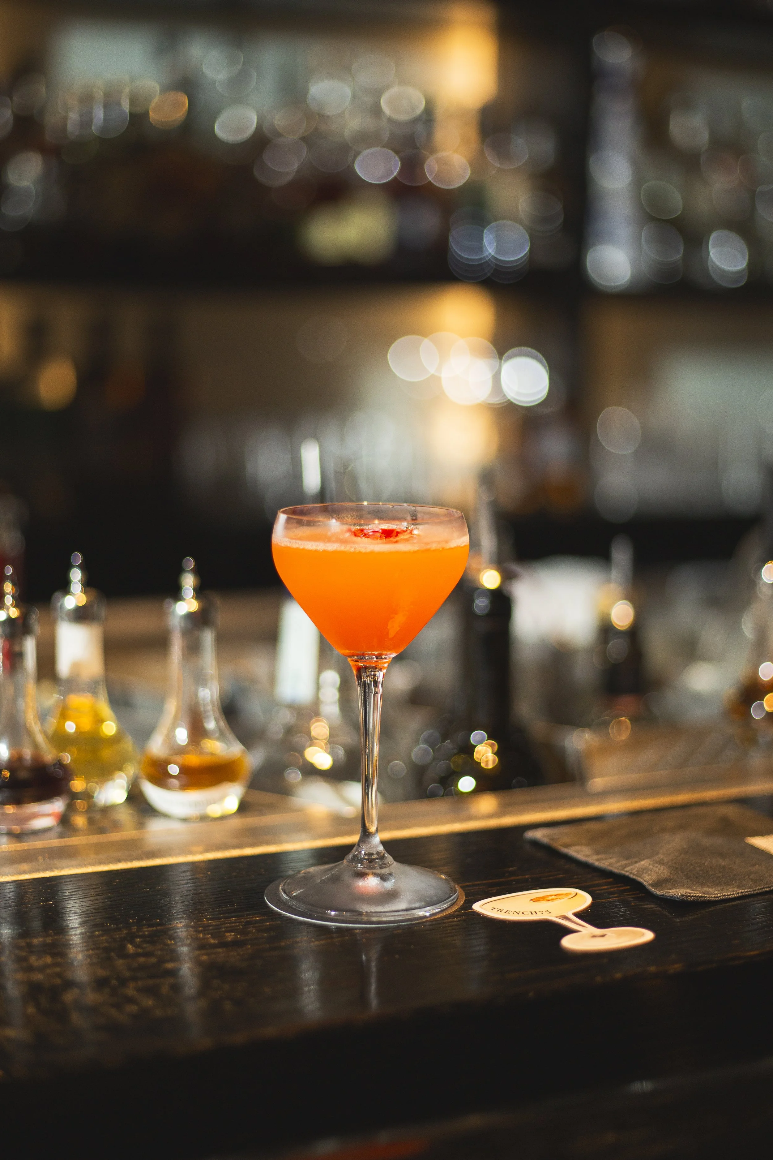 A pink cocktail in a coupe glass on a dark bar counter, with blurry bar bottles and shelves in the background.