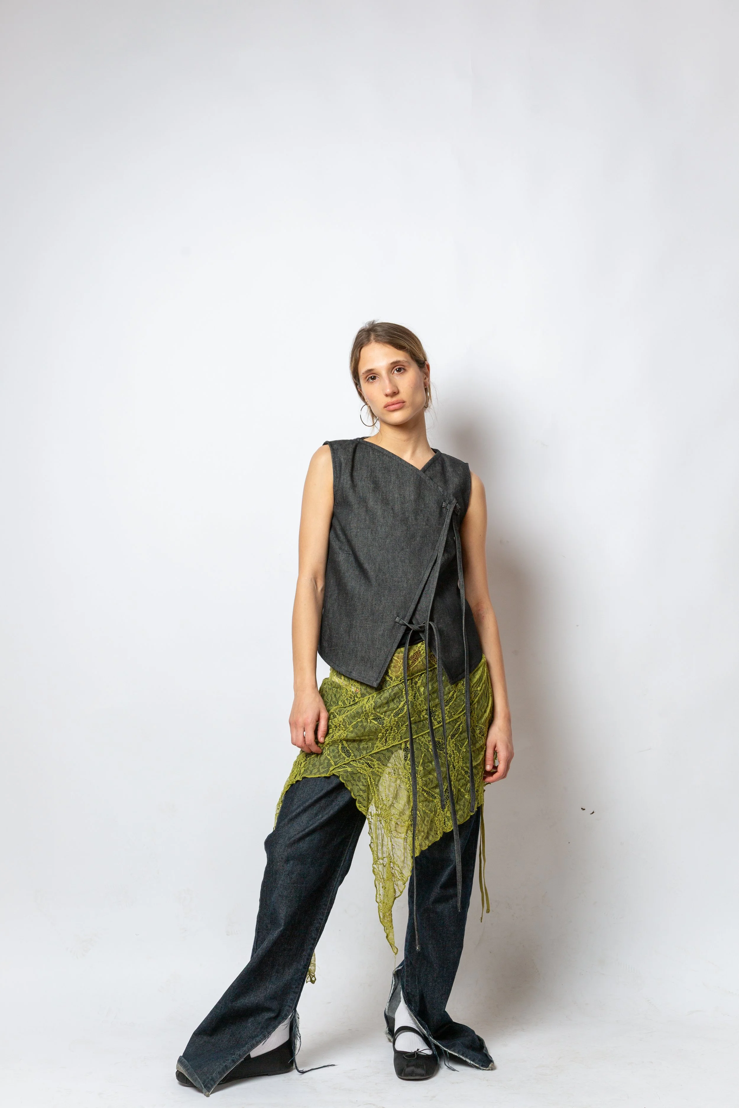 A young woman with brown hair styled in a loose bun, wearing a black sleeveless top, green lace detail skirt, and black oversized jeans, posing against a plain light-colored background.