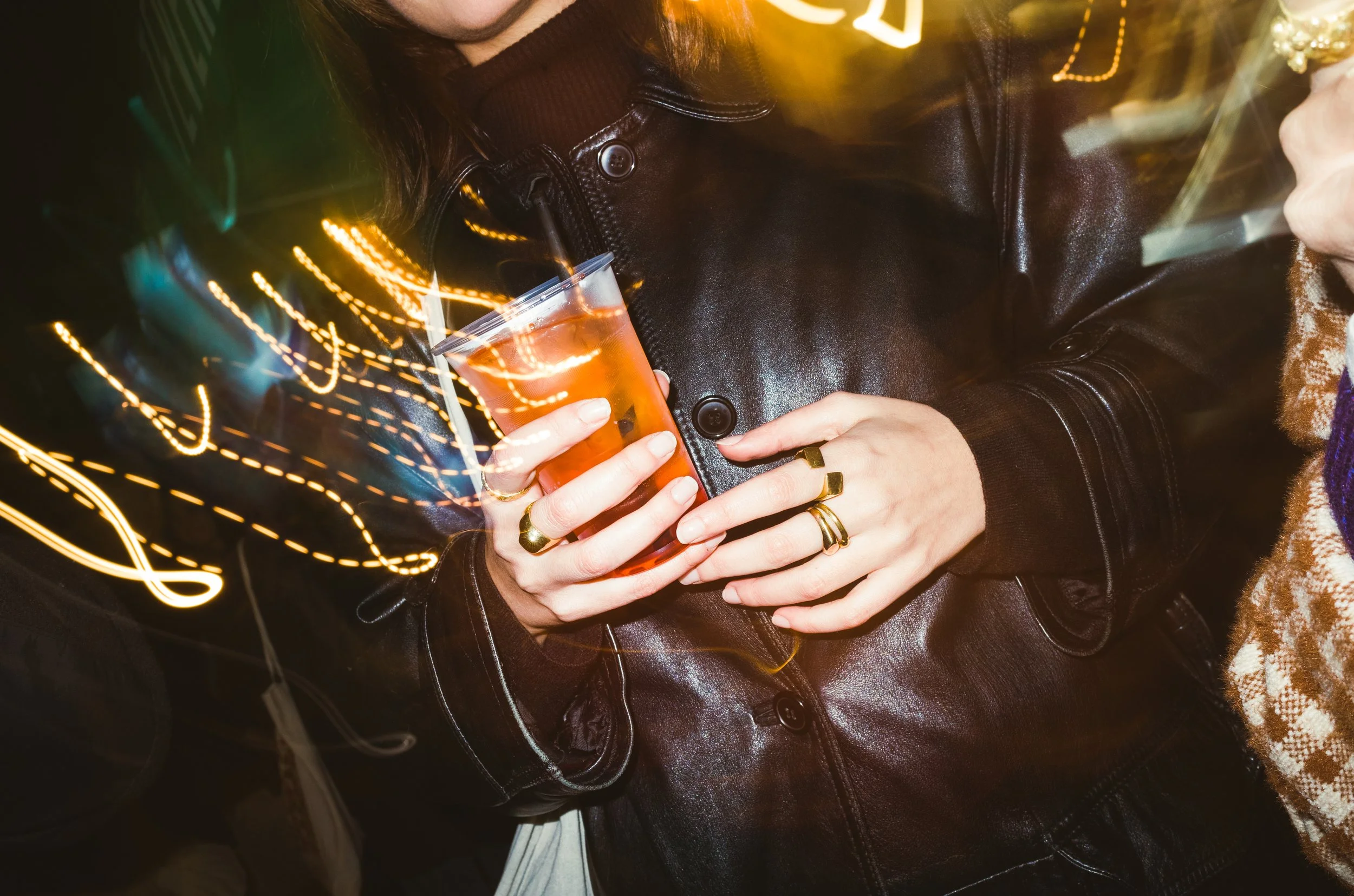 A person holding a plastic cup with a drink, wearing multiple rings and a black leather jacket, at a nighttime social event with colorful light streaks.