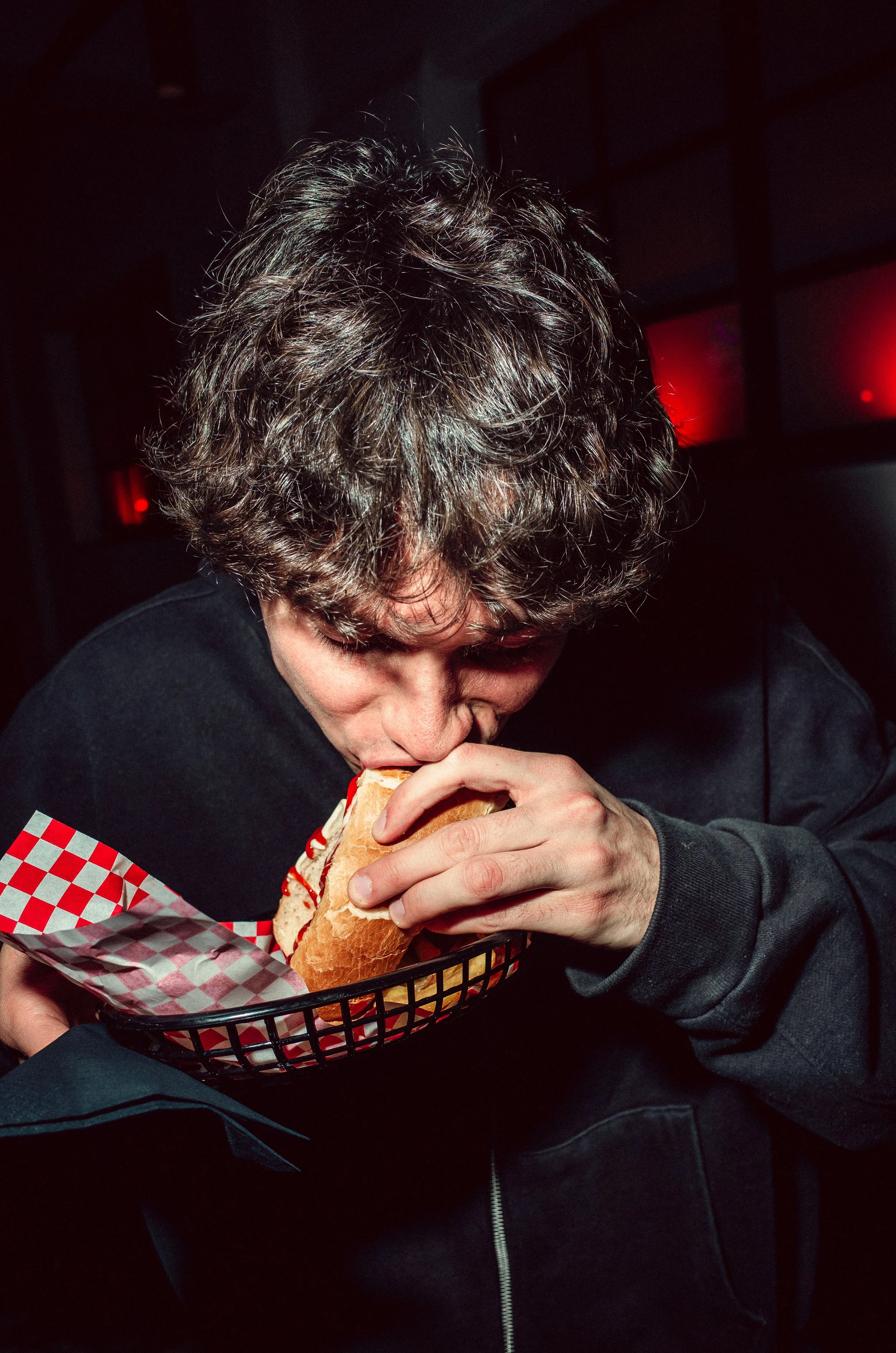 A person with curly hair is eating a hot dog in a basket with red and white checkered paper in a dimly lit environment.