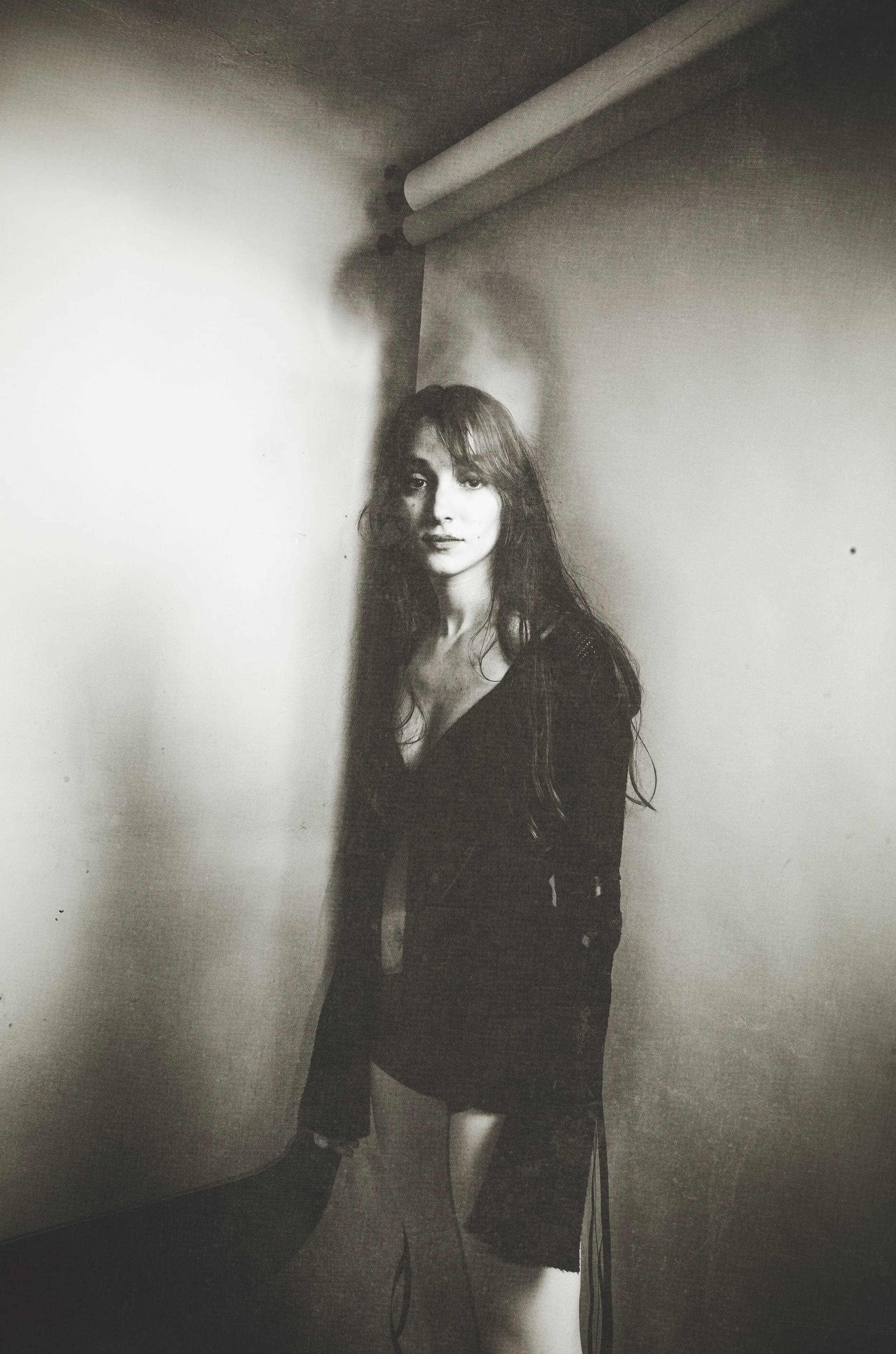 Black and white photo of a young woman with long hair, wearing a dark jacket and shorts, standing against a wall with a pipe above. She looks at the camera with a neutral expression.