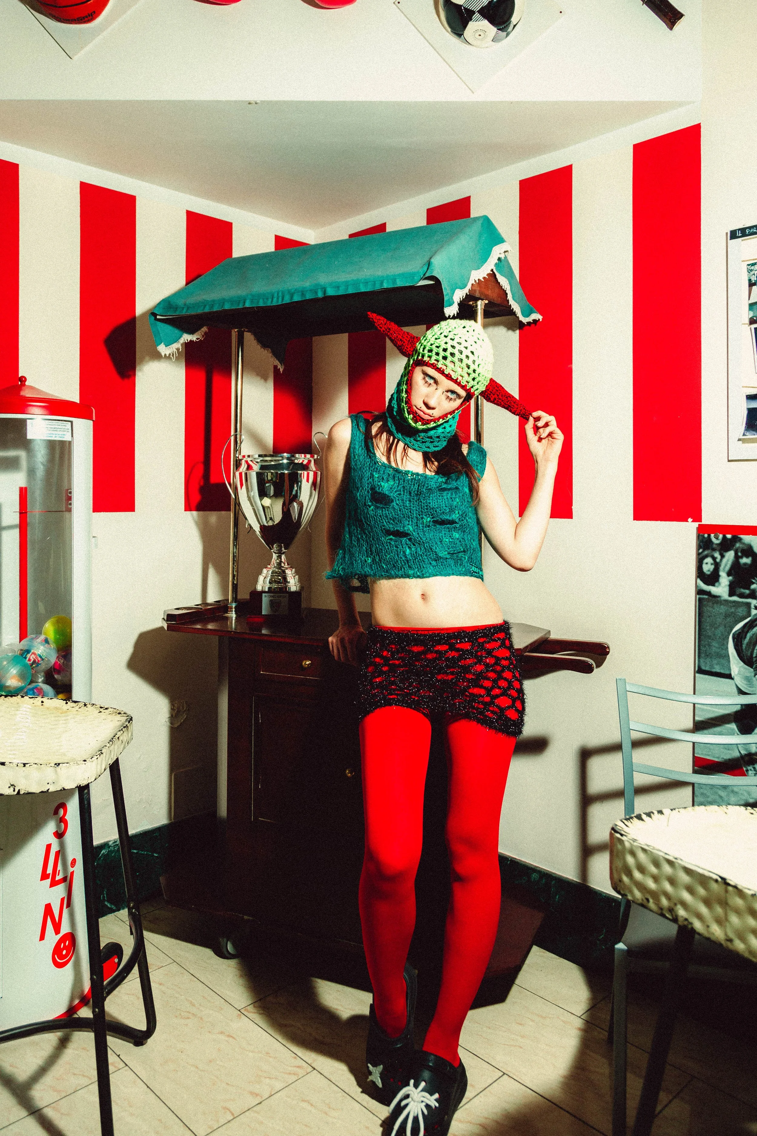 A young woman with red hair and tattoos wearing a green knit hat with earflaps and a red spike on top, a teal distressed sleeveless top, a black crochet skirt over red tights, and black sneakers with white laces, posing in a room with red and white s