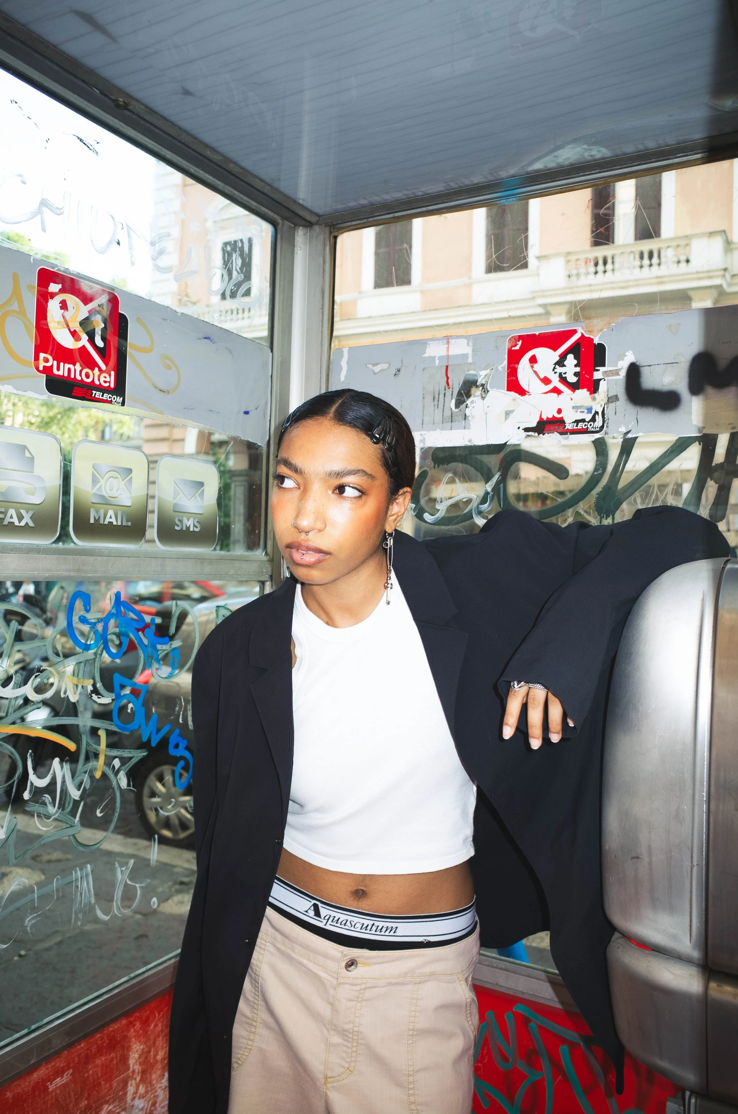 Young woman standing inside a phone booth, wearing a black blazer over a white crop top, beige pants, and silver jewelry, with graffiti on the glass window.