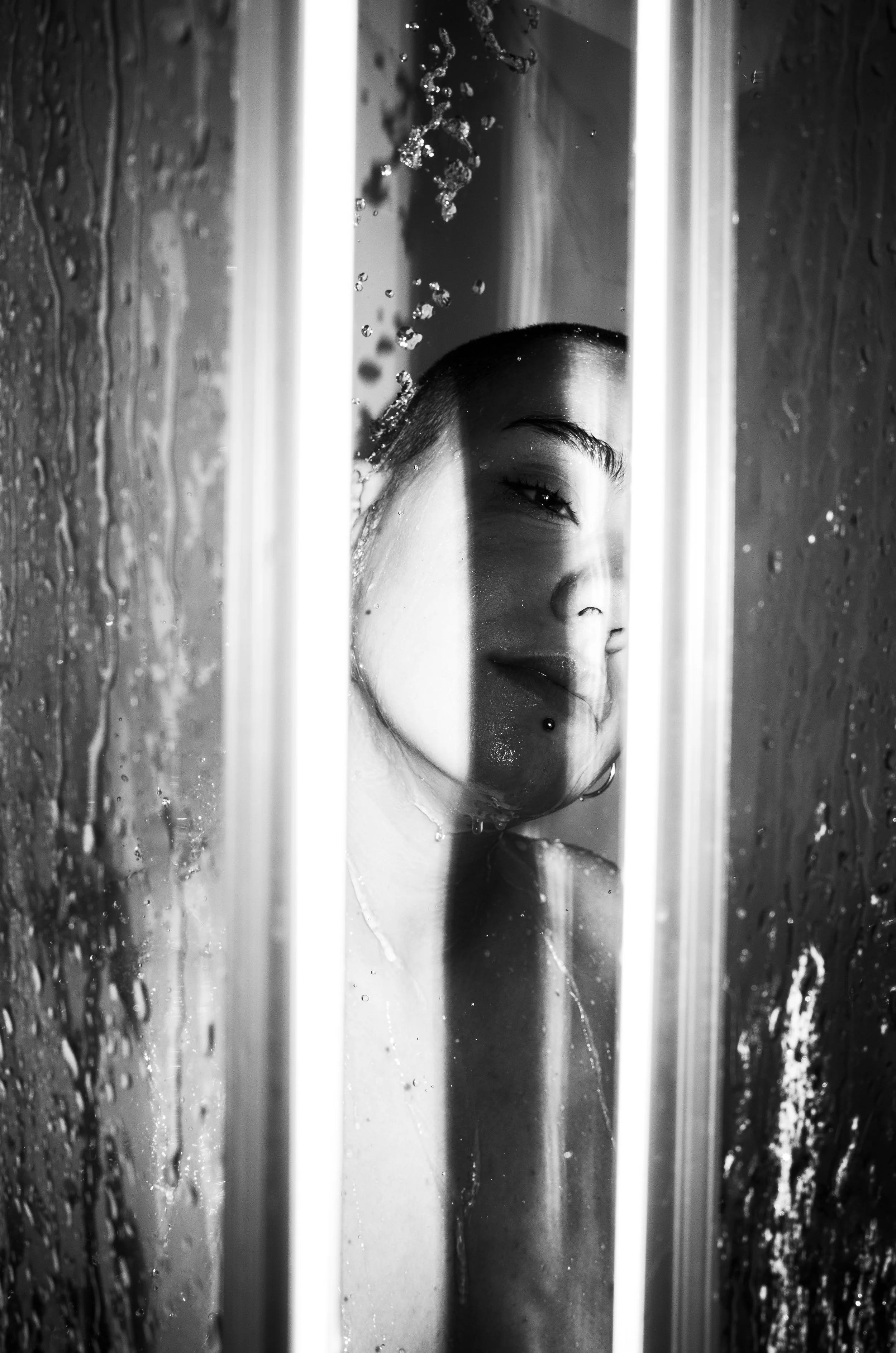 A black and white photo of a woman taking a shower, with water falling on her face and a partial view of her face visible through a glass shower door.