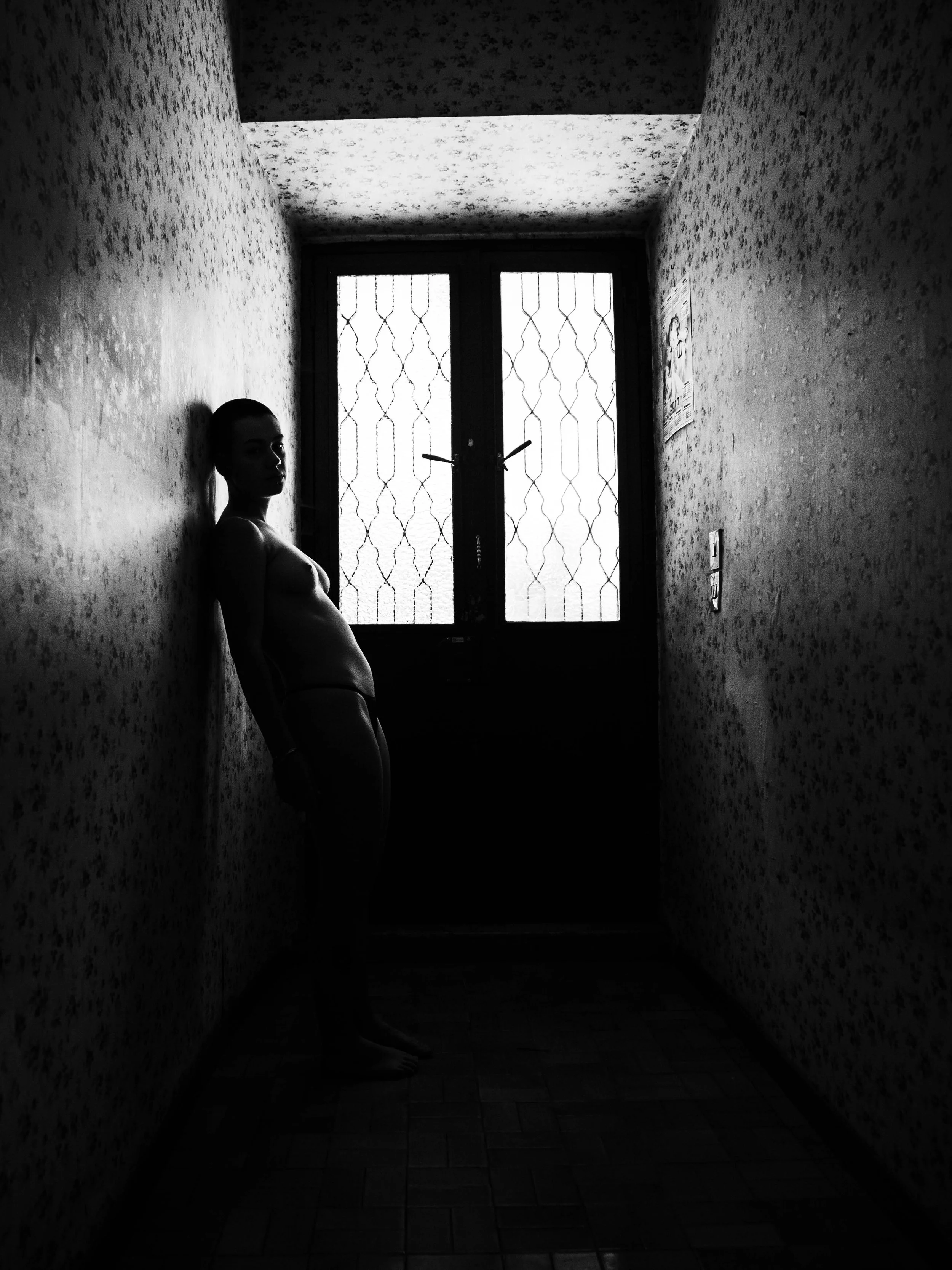 Silhouette of a woman standing in a narrow hallway, backlit by a window with decorative glass, creating a dark contrast in a black and white photo.