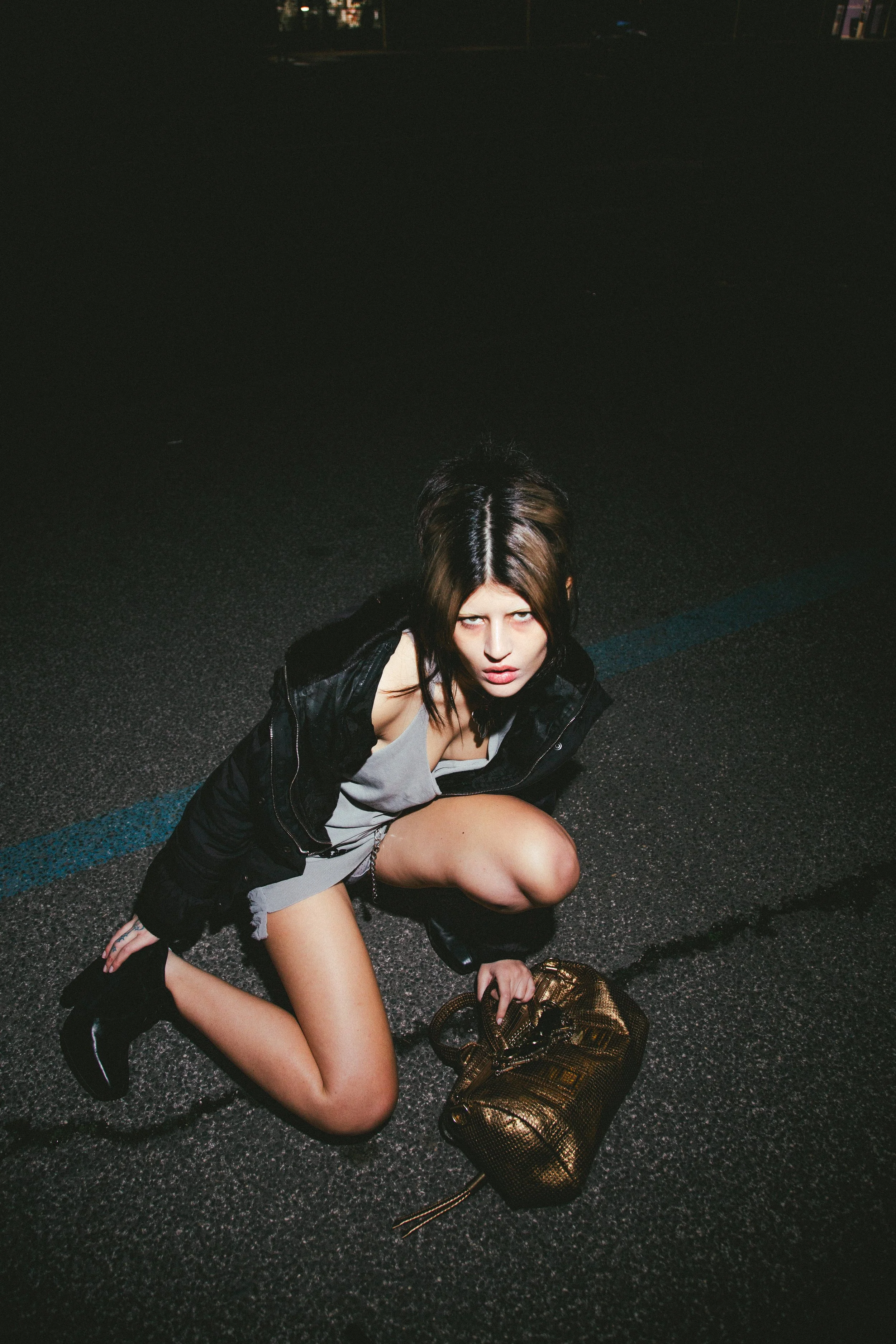 A woman crouching on a dark street at night, holding a gold handbag, wearing a black jacket, gray dress, and black boots, with an intense expression.