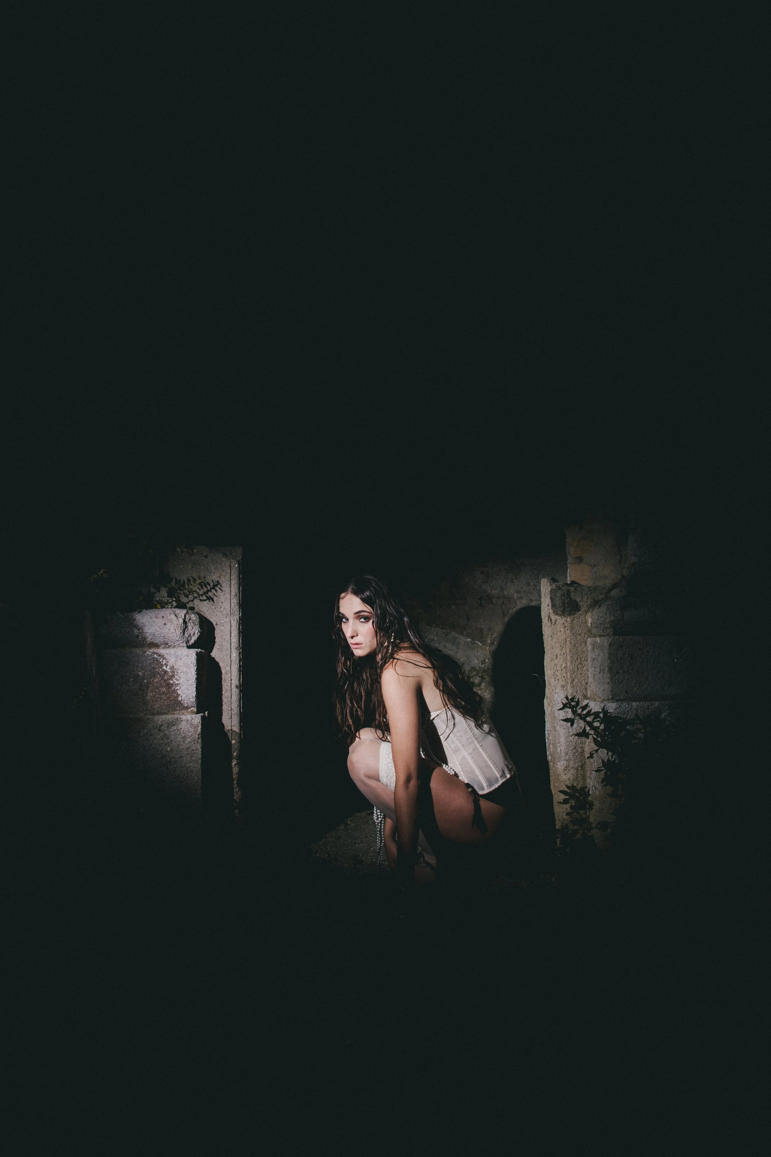 A woman with long dark hair crouches outdoors near stone ruins or steps, illuminated by a focused light, with a dark background.