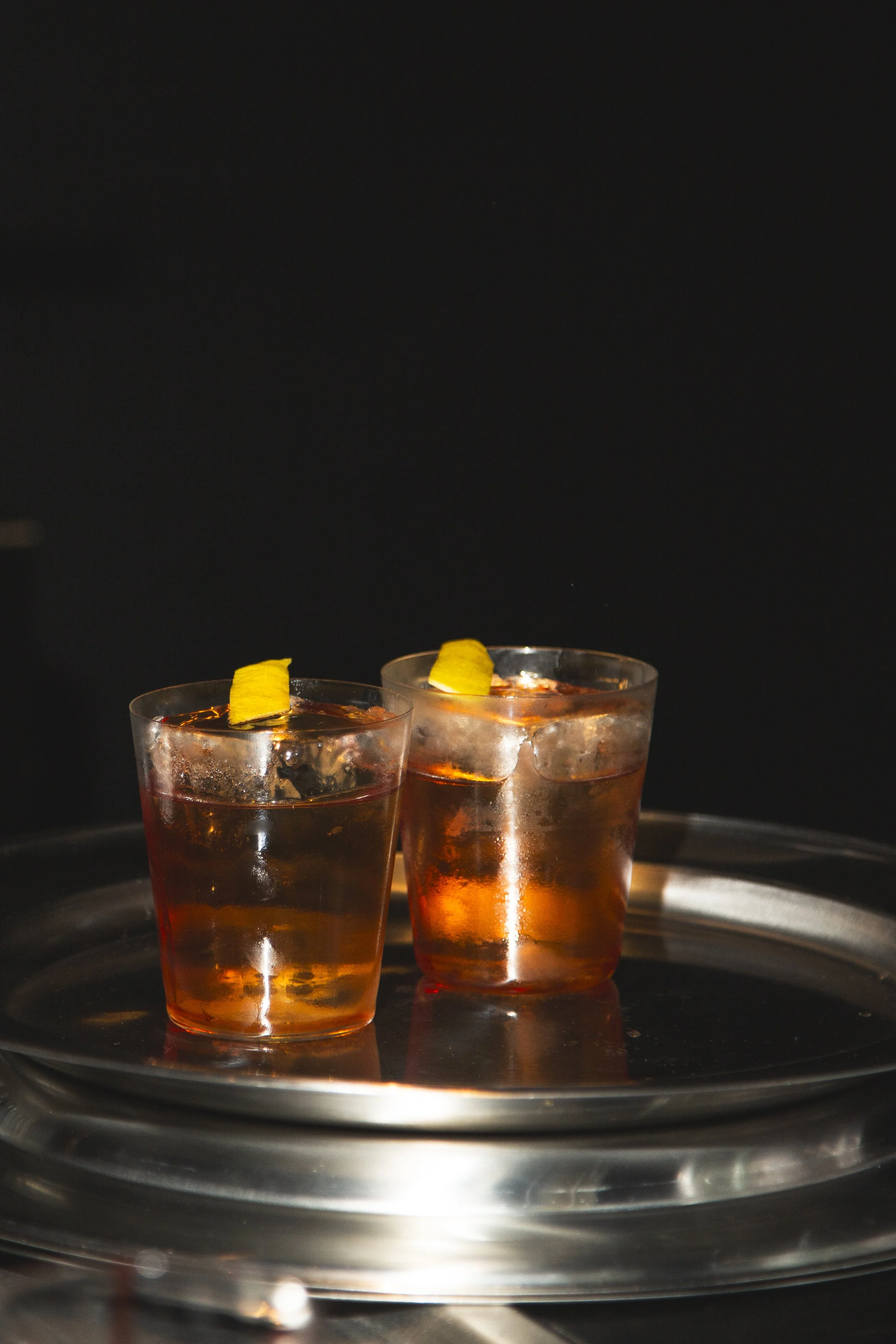 Two glasses of dark-colored cocktail with lemon wedges, placed on a silver tray against a black background.