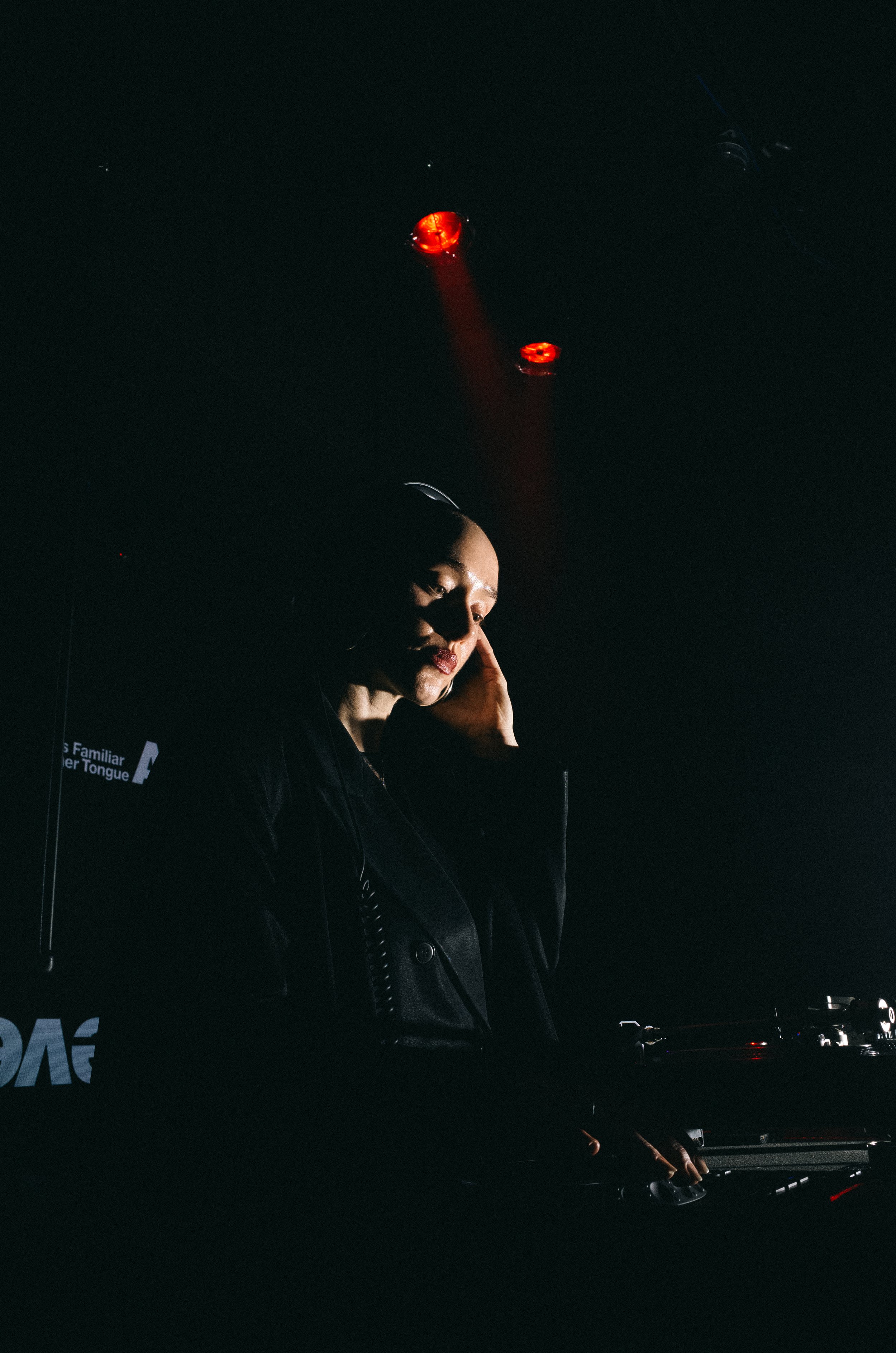 A female DJ wearing headphones, standing in a dark room with red and blue lighting, adjusting her equipment.
