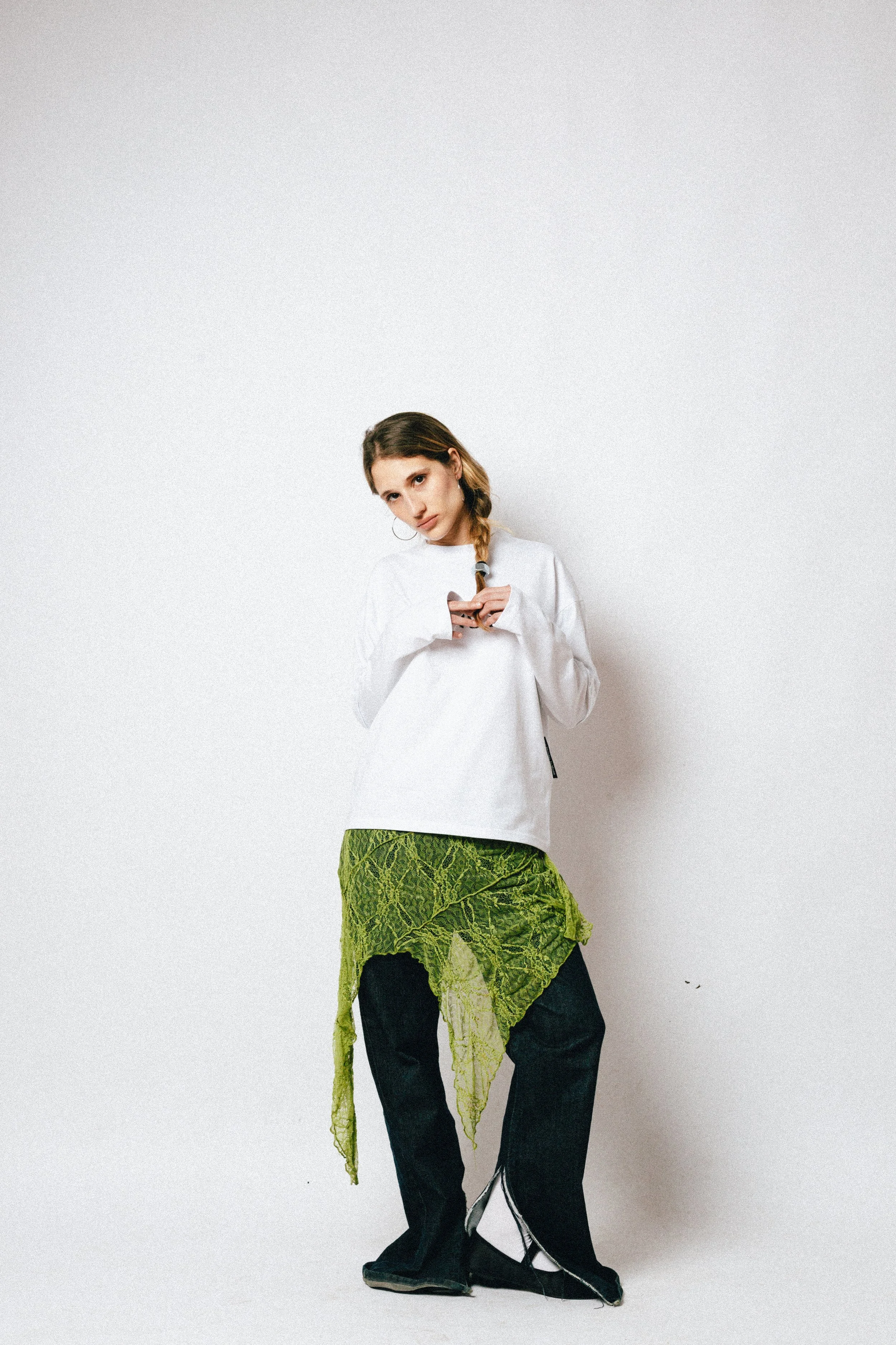 A young woman with light brown hair styled in a braid, wearing a white sweatshirt, black pants, and a green textured fabric draped around her waist. She stands against a plain white background, holding a small object, and looks directly at the camera with a neutral expression.