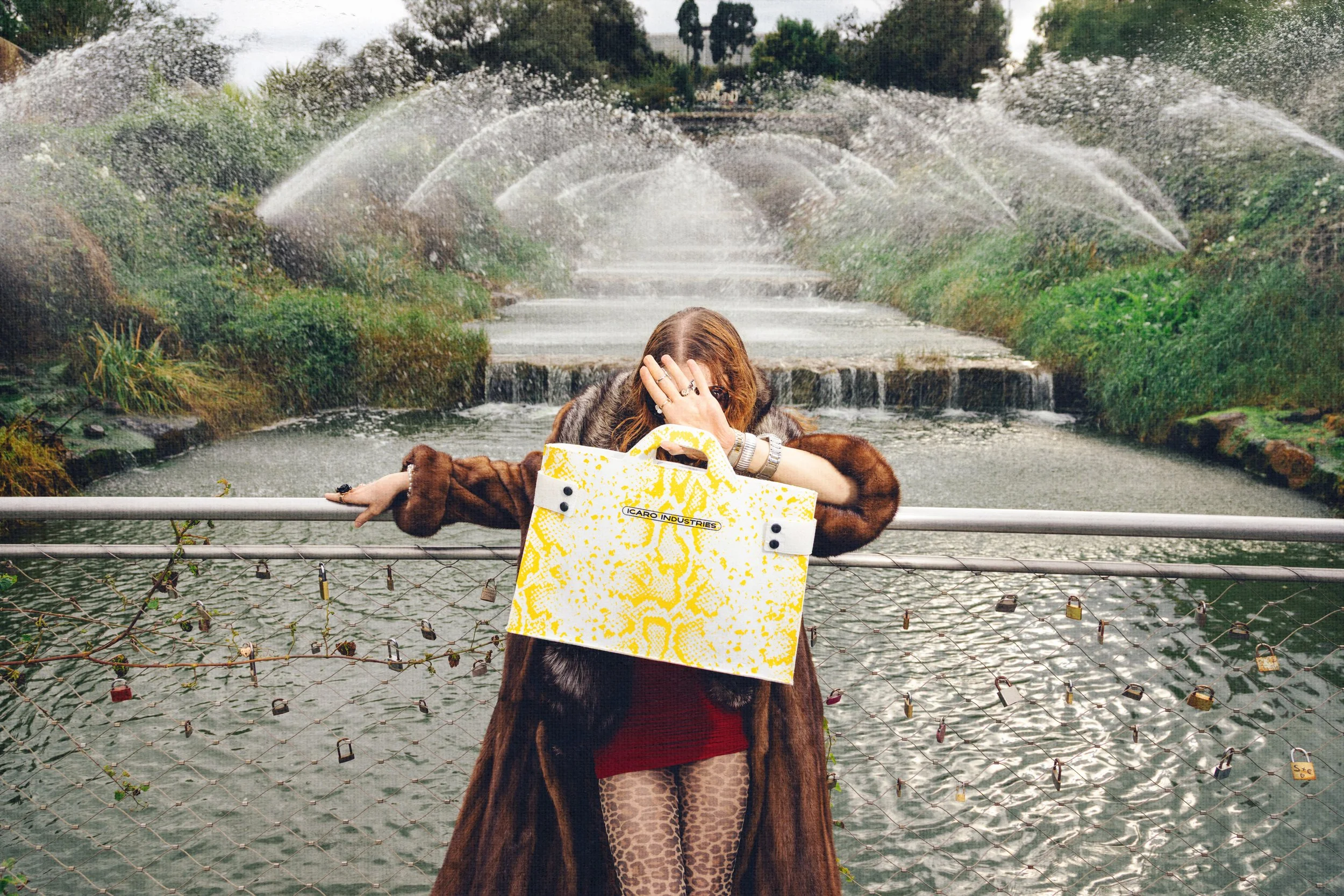 A woman standing in front of a water feature with spraying water fountains, holding a yellow patterned handbag in front of her face, wearing a brown fur coat and leopard print pants.