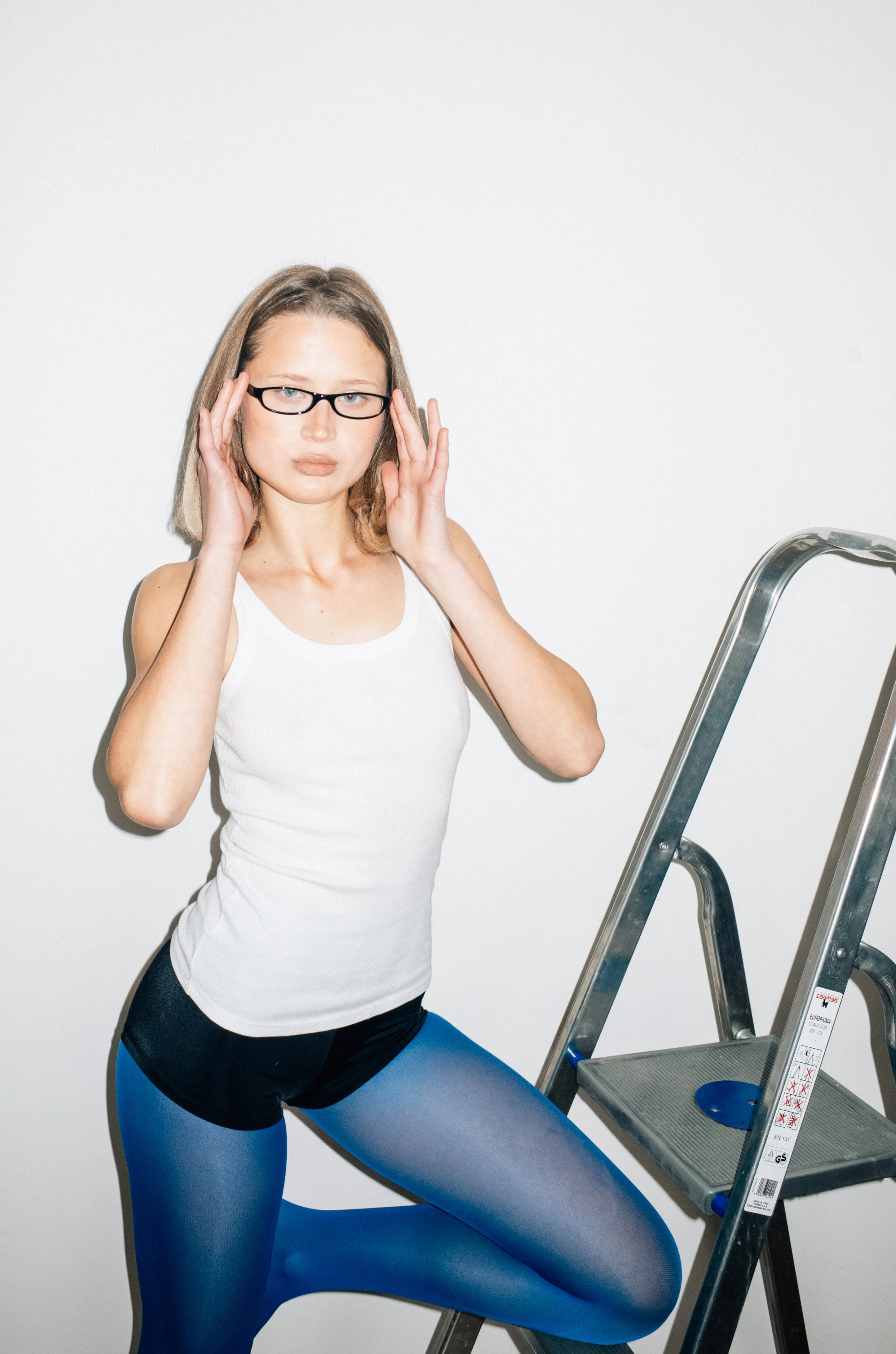 A woman with glasses in a white tank top and blue leggings standing next to a step ladder, adjusting her glasses and looking at the camera.
