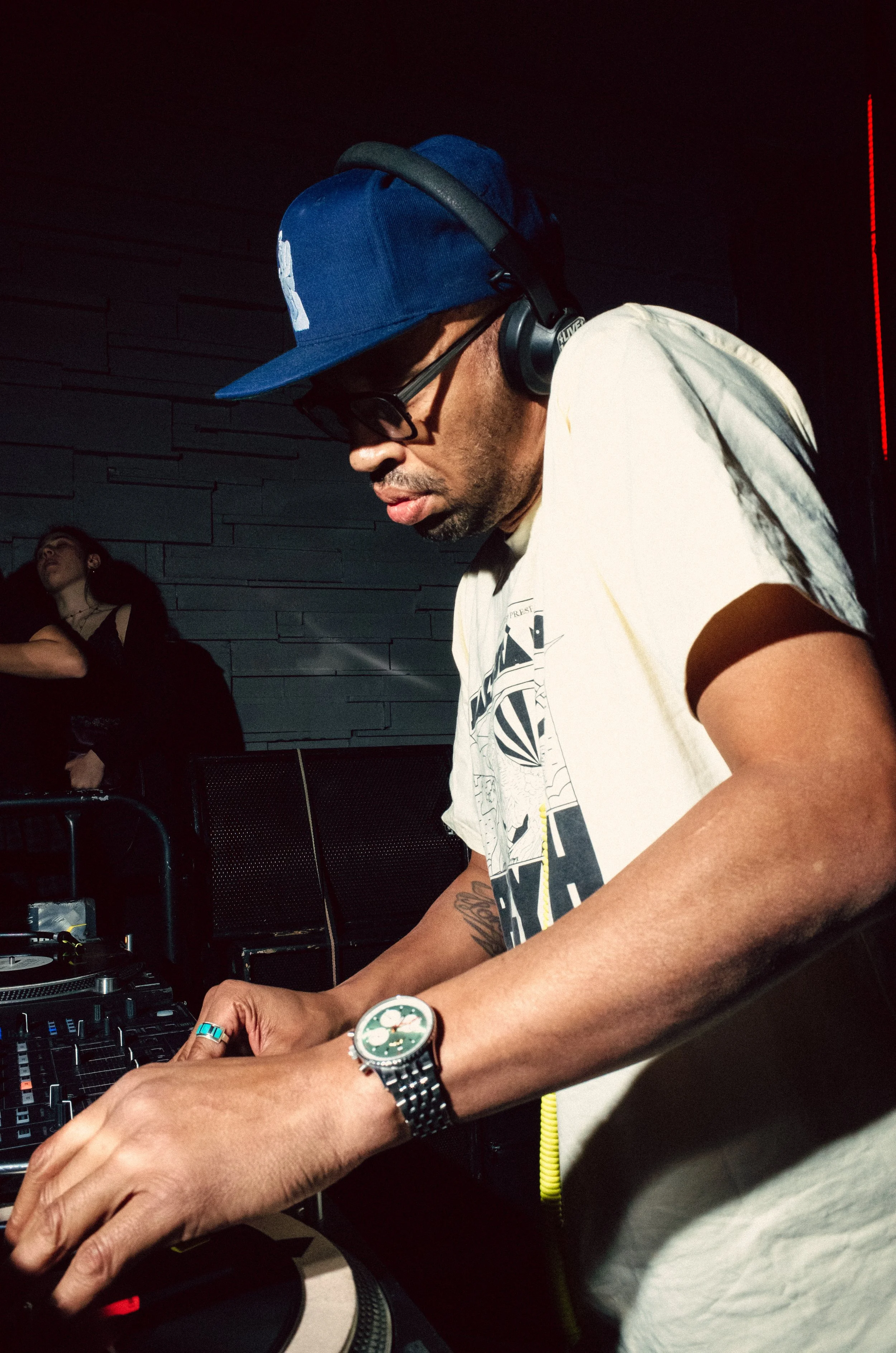 A male DJ wearing a blue cap, glasses, and headphones, mixing music at a nightclub or party.