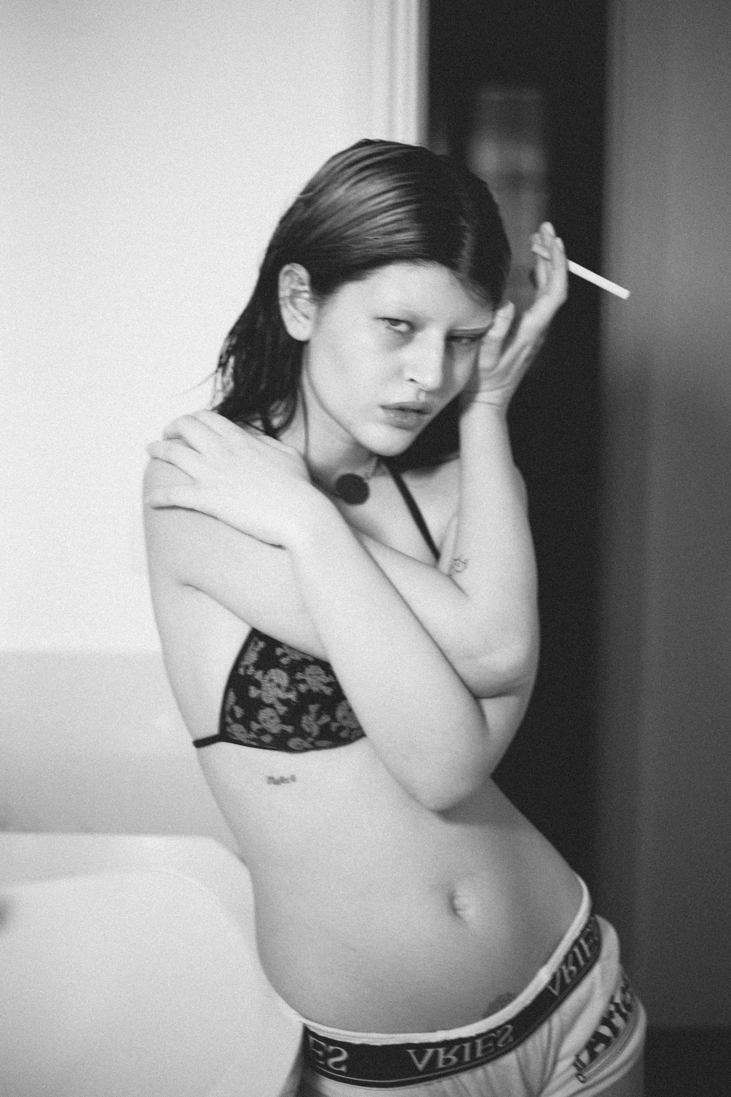 A black and white photo of a young woman with dark hair, wearing a floral bra and shorts, holding a cigarette, and posing with an intense expression.