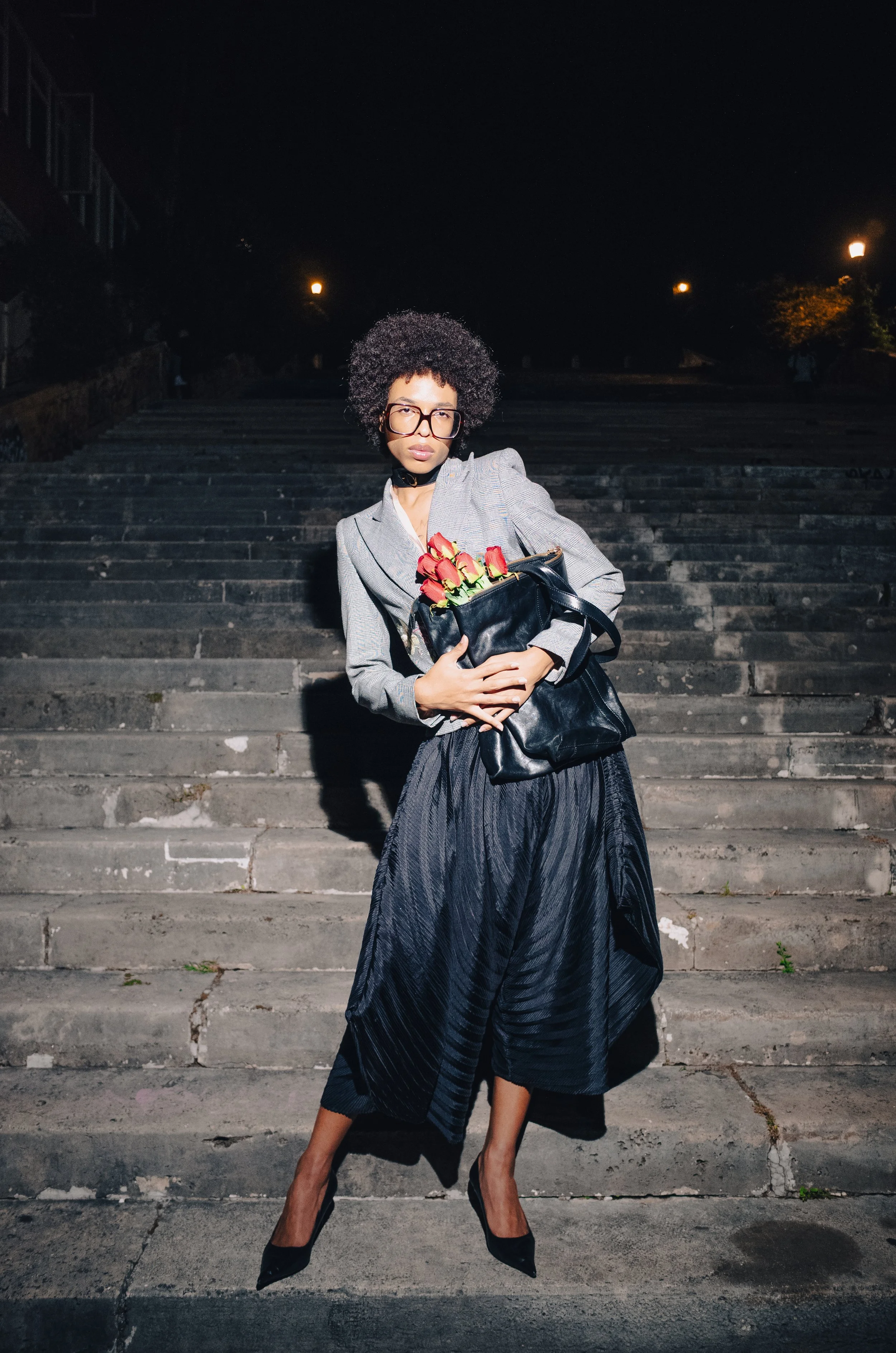 A woman standing on concrete stairs at night, wearing a gray blazer, black pleated skirt, and black high heels, holding a black purse with red roses inside, with a serious expression and glasses.