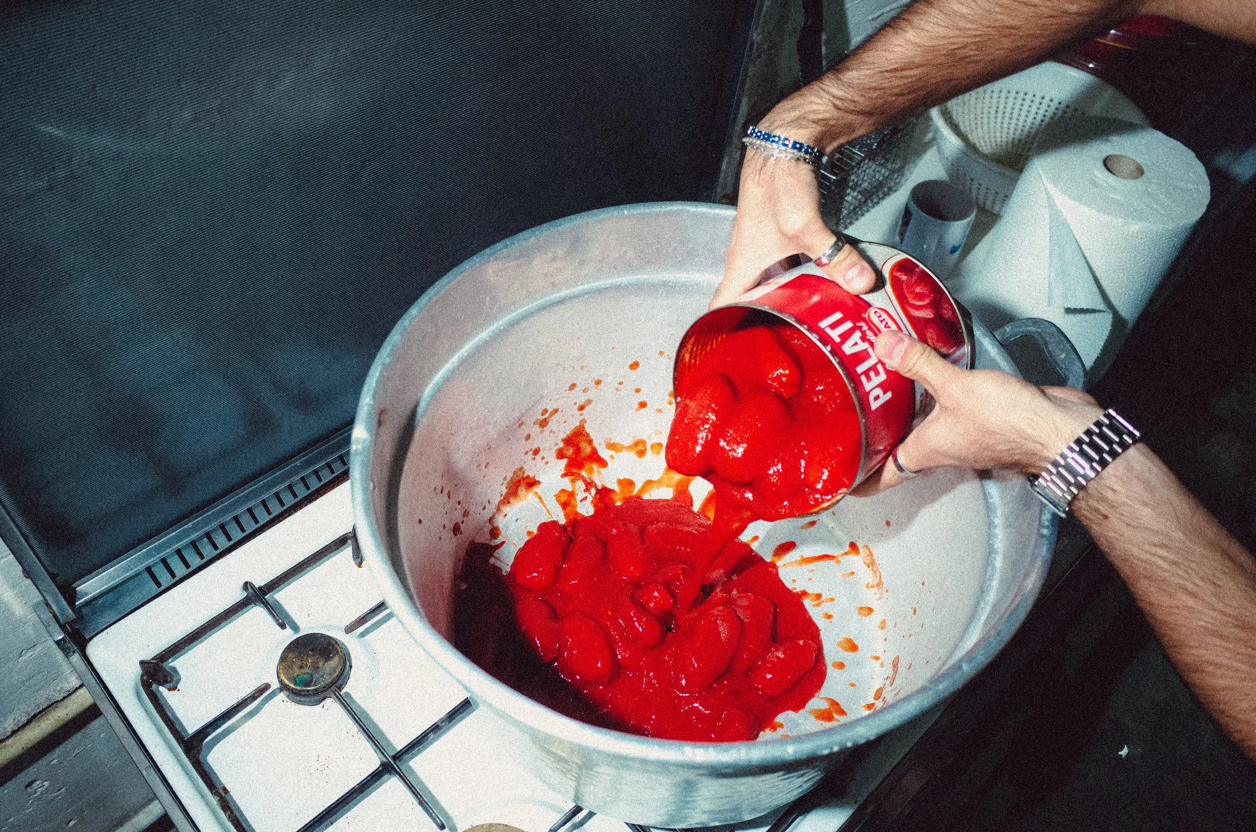 Person pouring canned tomato sauce into a large pot on a stove.