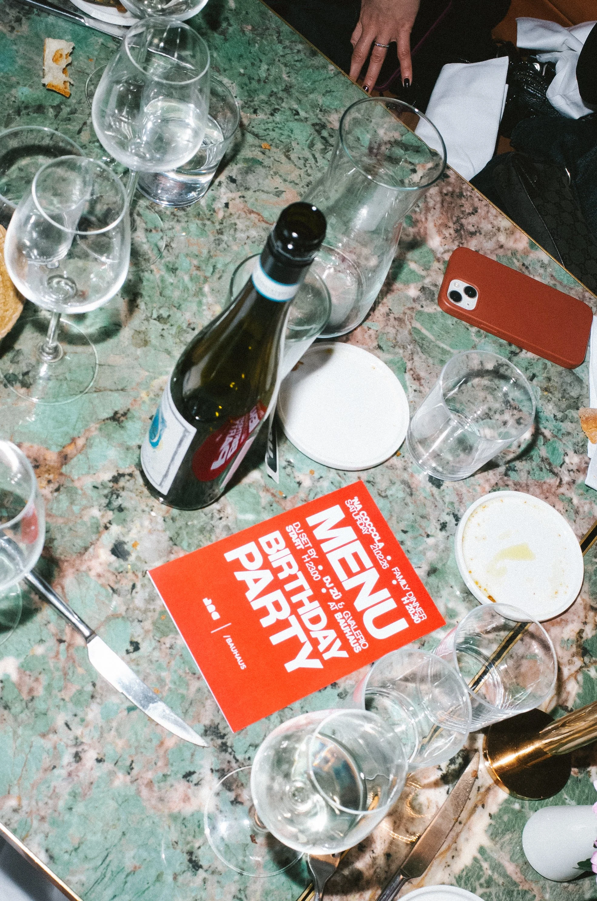 A table with empty wine glasses, a beer bottle, a smartphone, a plate with leftover food, a piece of bread, and a flyer for a birthday party.