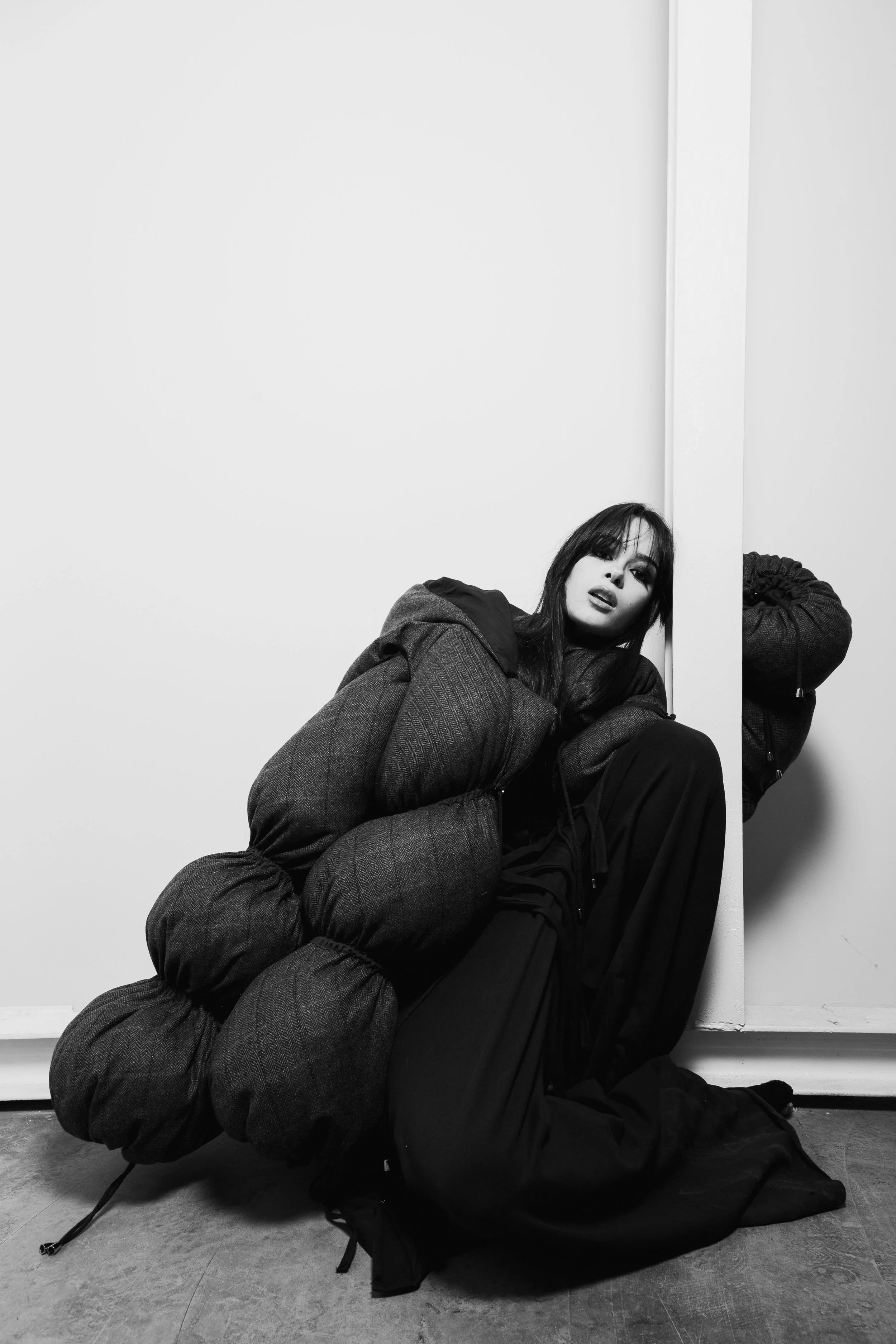 A black and white photo of a woman sitting on the floor with a large, puffy, padded coat wrapped around her. She has dark hair, is wearing dark clothing, and poses against a plain background with a corner wall. She looks at the camera with a serious 