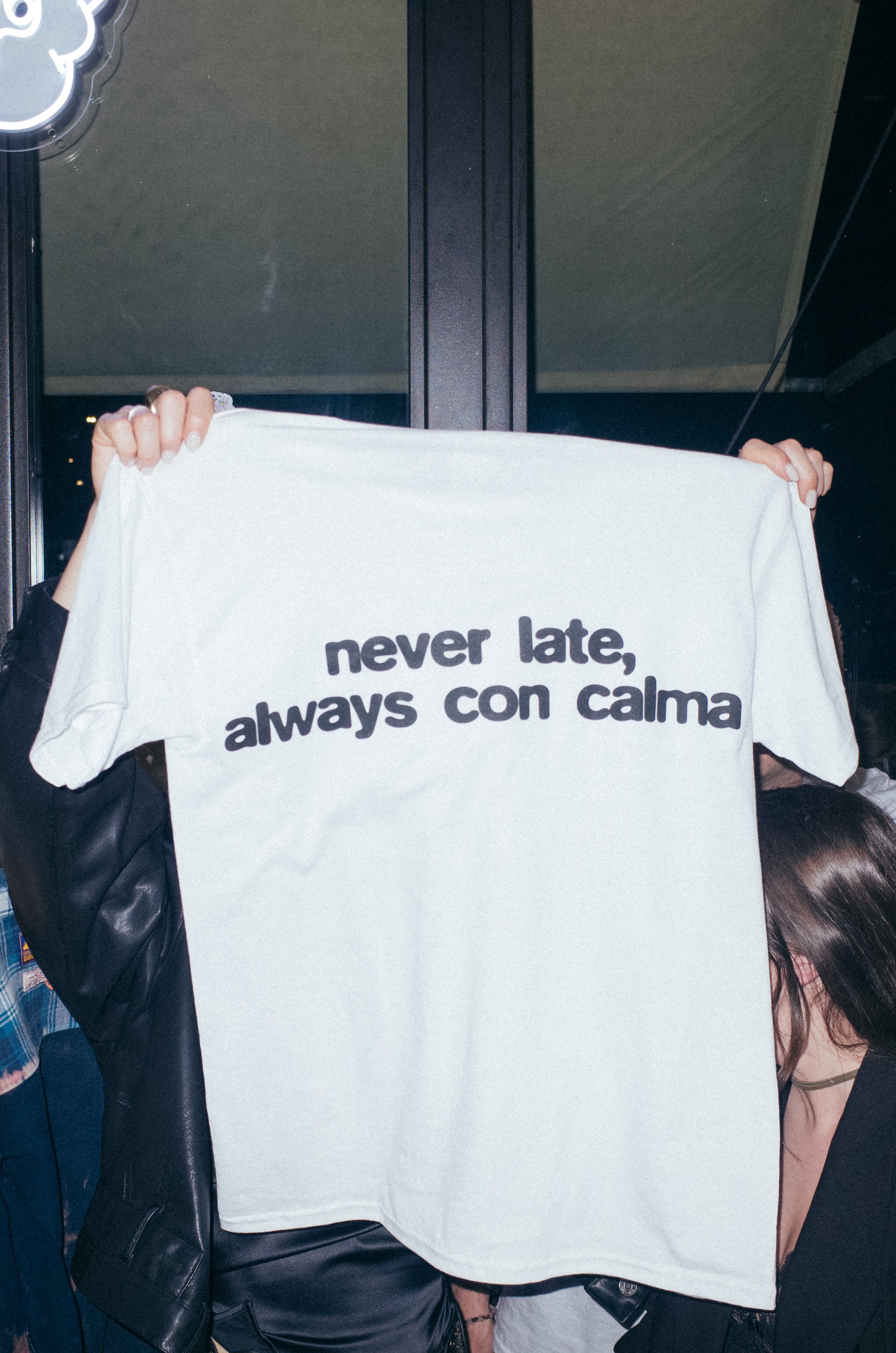 Person holding a white T-shirt with the text "never late, always con calma" written on it, partially obscuring their face.