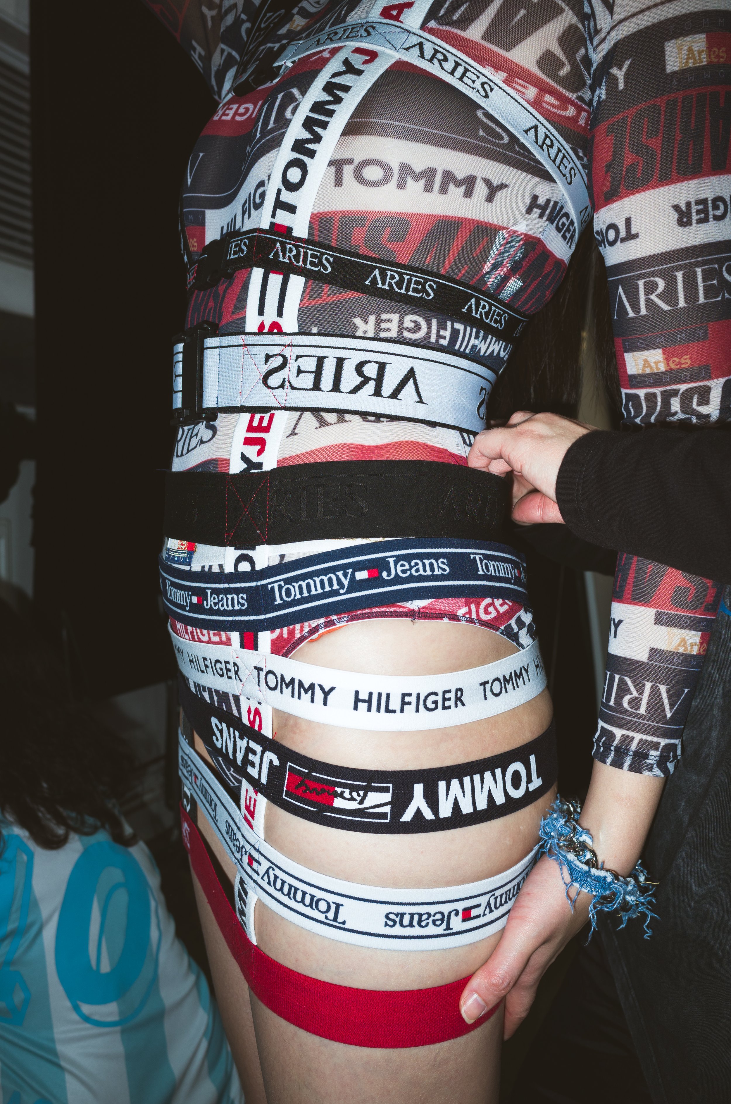 Person wearing multiple Tommy Hilfiger and Tommy Jeans branded straps around waist and thigh, with colorful clothing featuring brand logos.