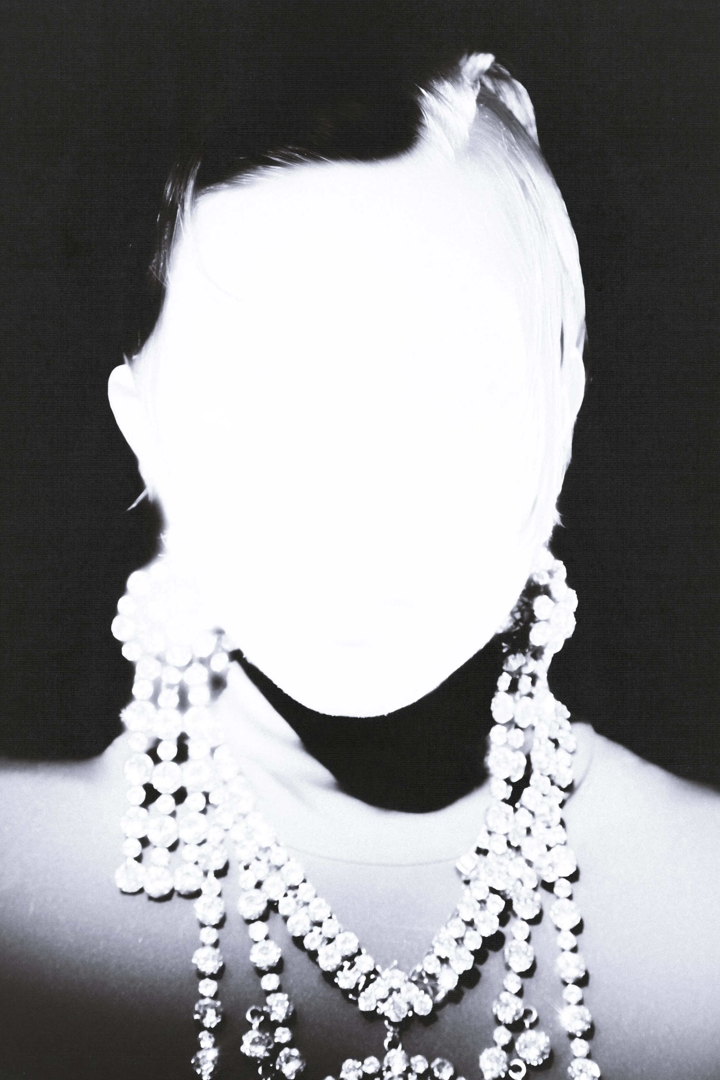 Black and white photograph of a faceless person wearing multiple strands of large, shiny necklaces. The person's face is overexposed or erased, and they have short, dark hair and earrings.