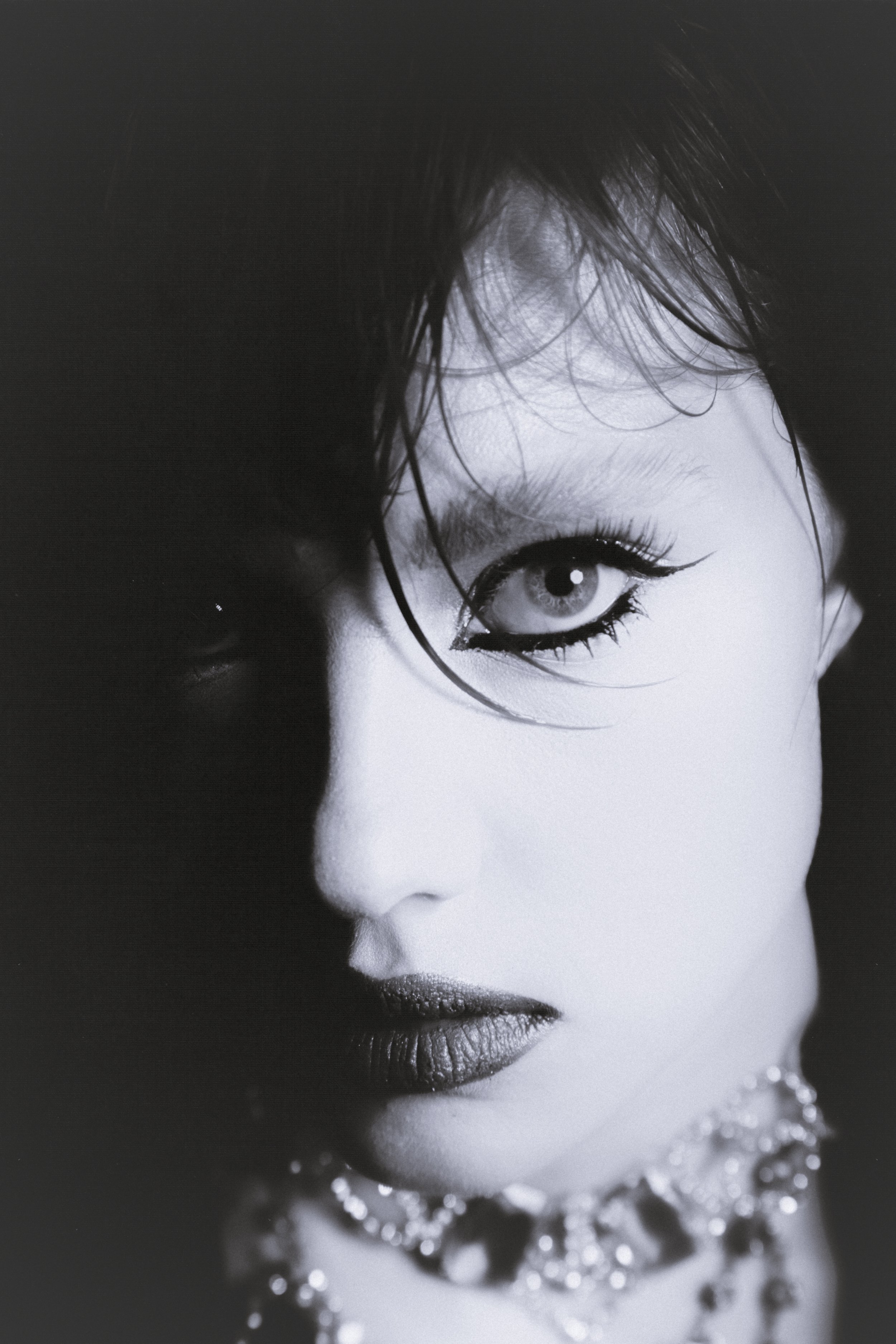 A black-and-white close-up portrait of a woman with dark, wet hair, striking eye makeup, and a necklace.