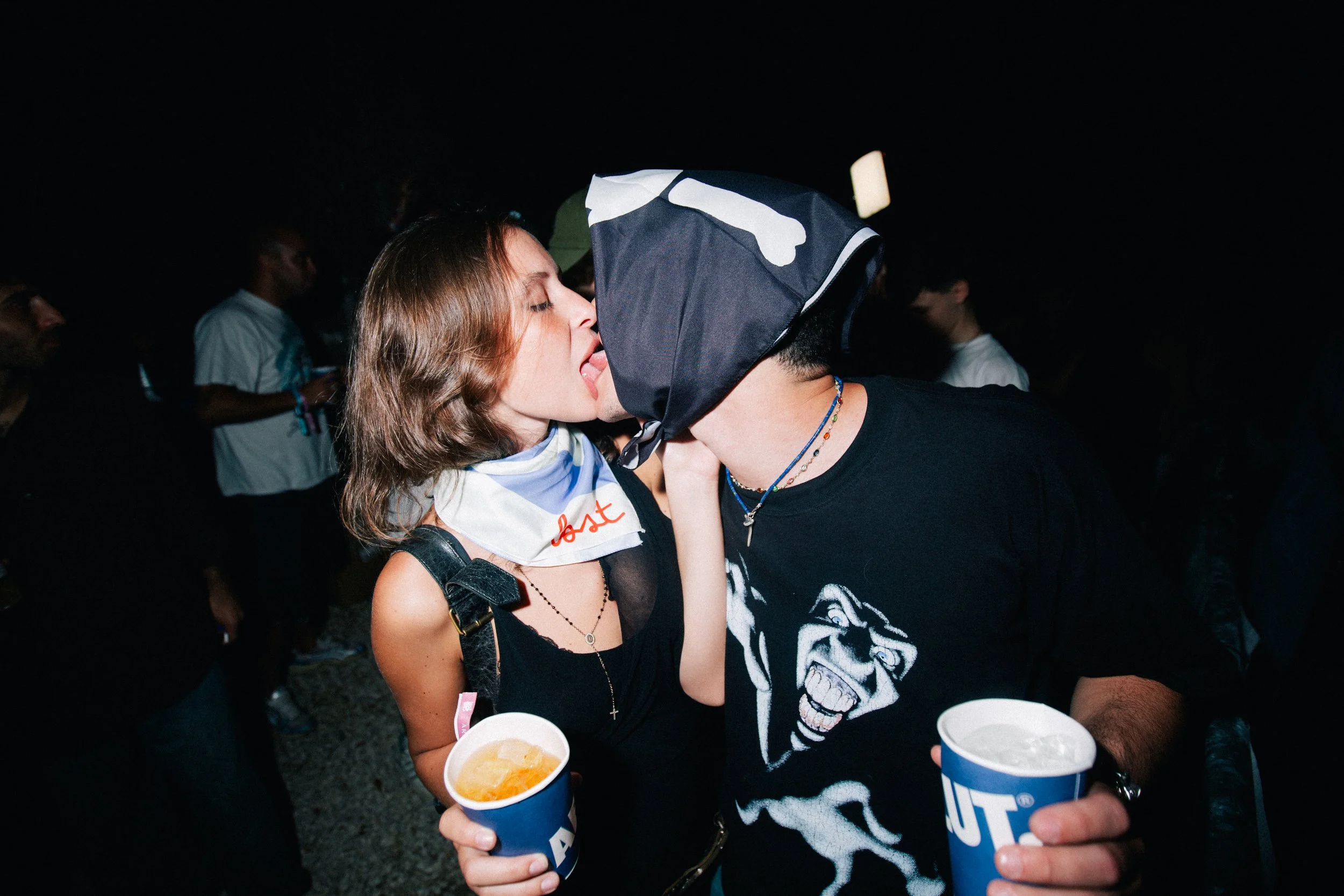 A young woman and a man are kissing at a nighttime outdoor event. The woman is wearing a black top, a necklace, and a scarf around her neck. The man is wearing a black T-shirt with a graphic of a snarling animal and a head covering. Both are holding 
