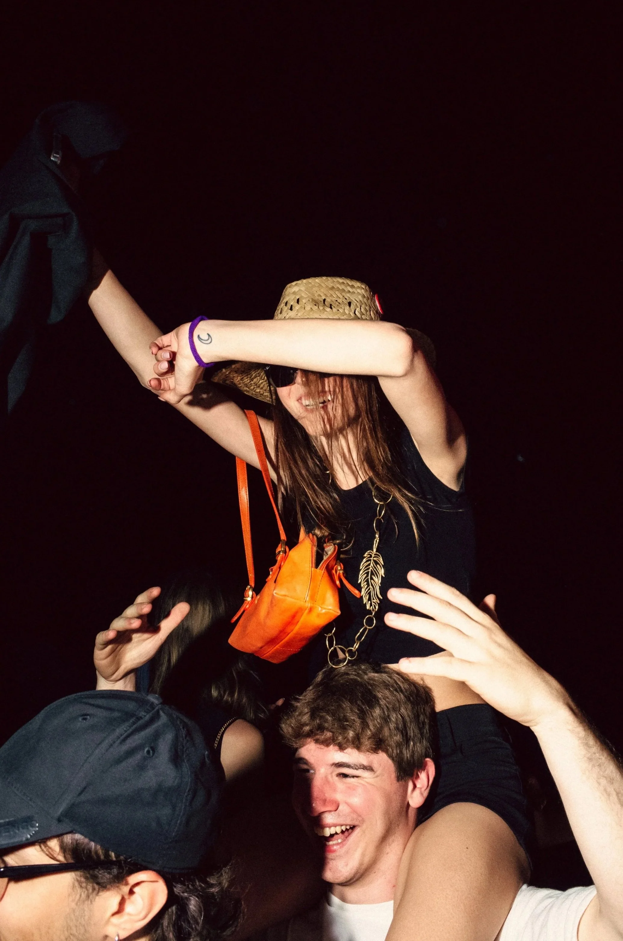 Group of young people celebrating at a party, one woman being lifted by a man, wearing sunglasses, hat, and a black tank top, smiling with her arm covering her eyes, friends enjoying in a dark setting.