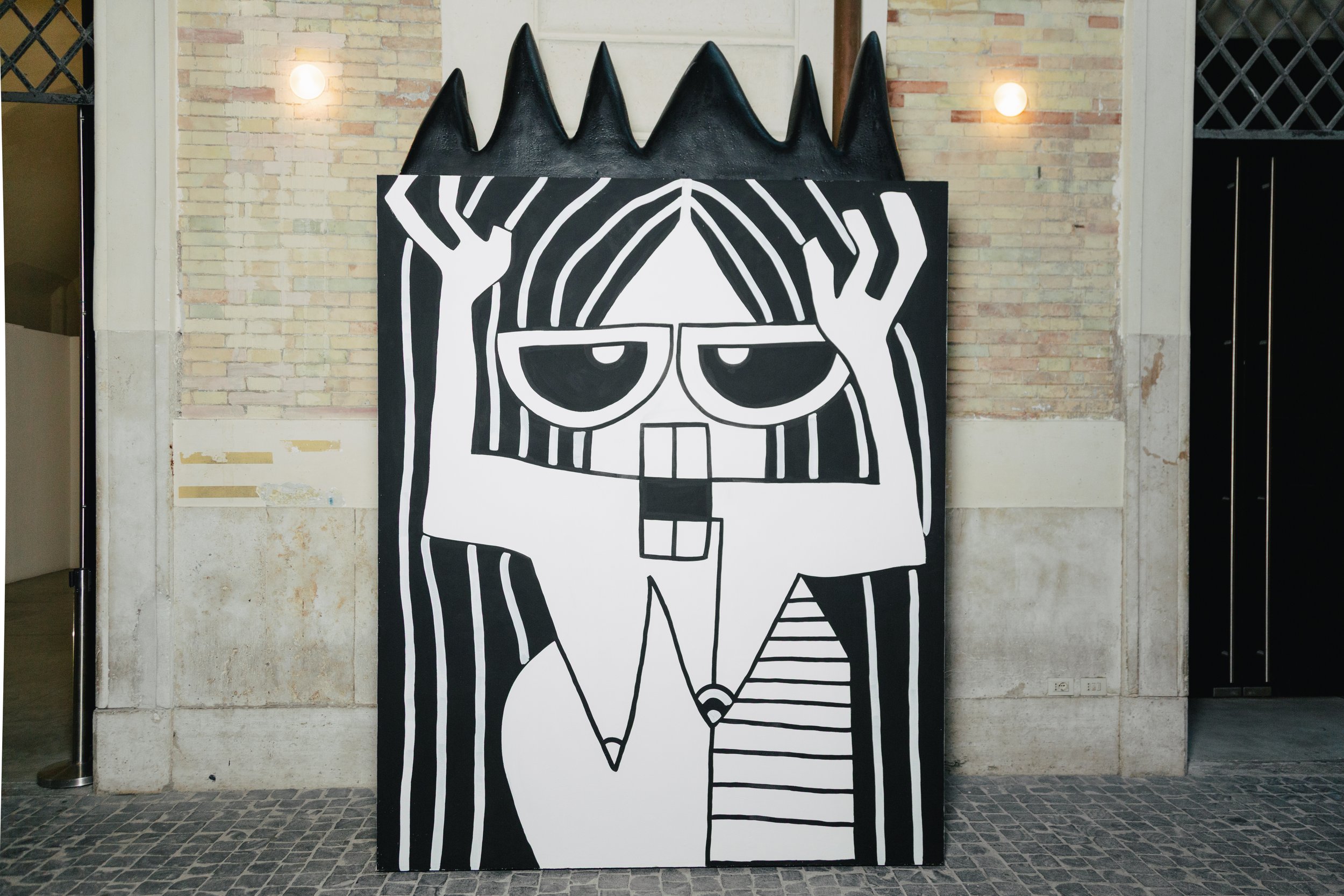 Black and white abstract art of a cartoon character with glasses and spiky hair, holding its head with hands, painted on a large rectangular canvas leaning against a brick wall.