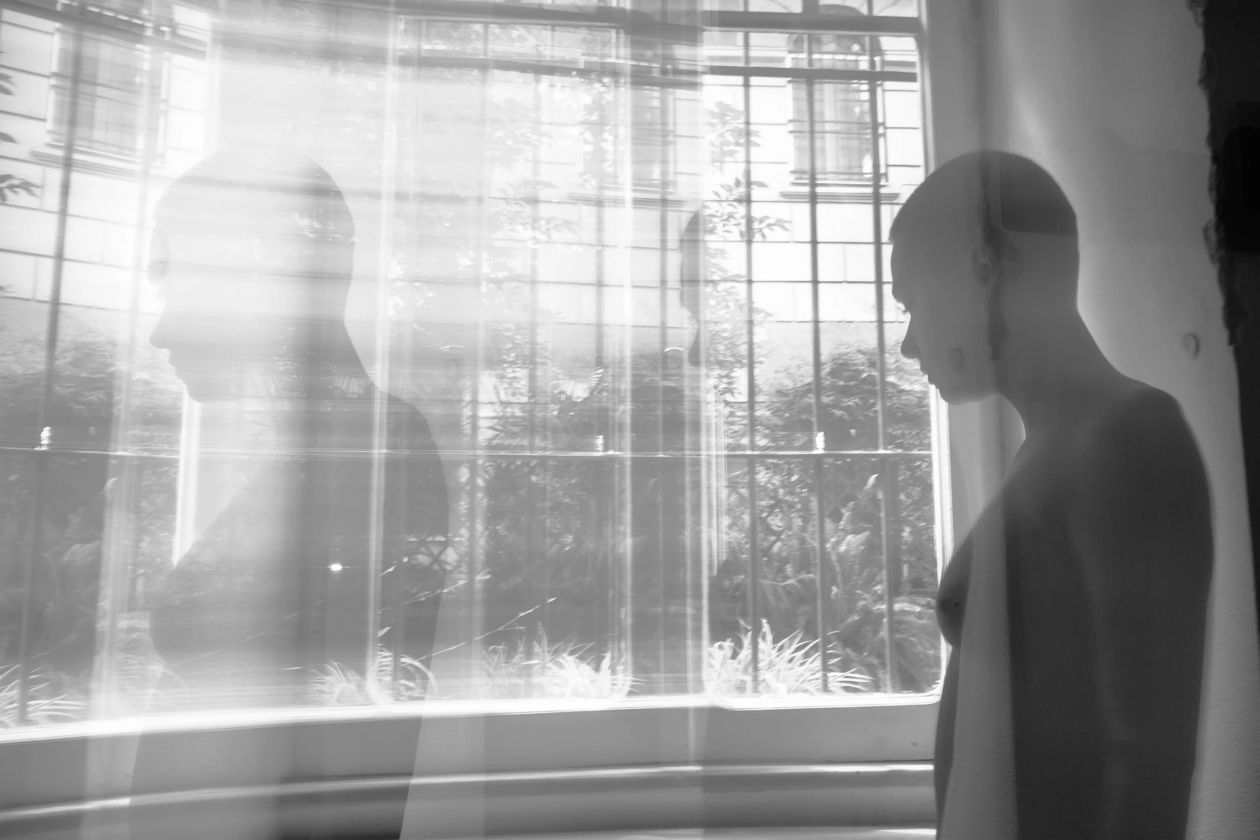 A black and white photo of a woman and a mannequin or reflection in front of a window with blinds, with trees visible outside.