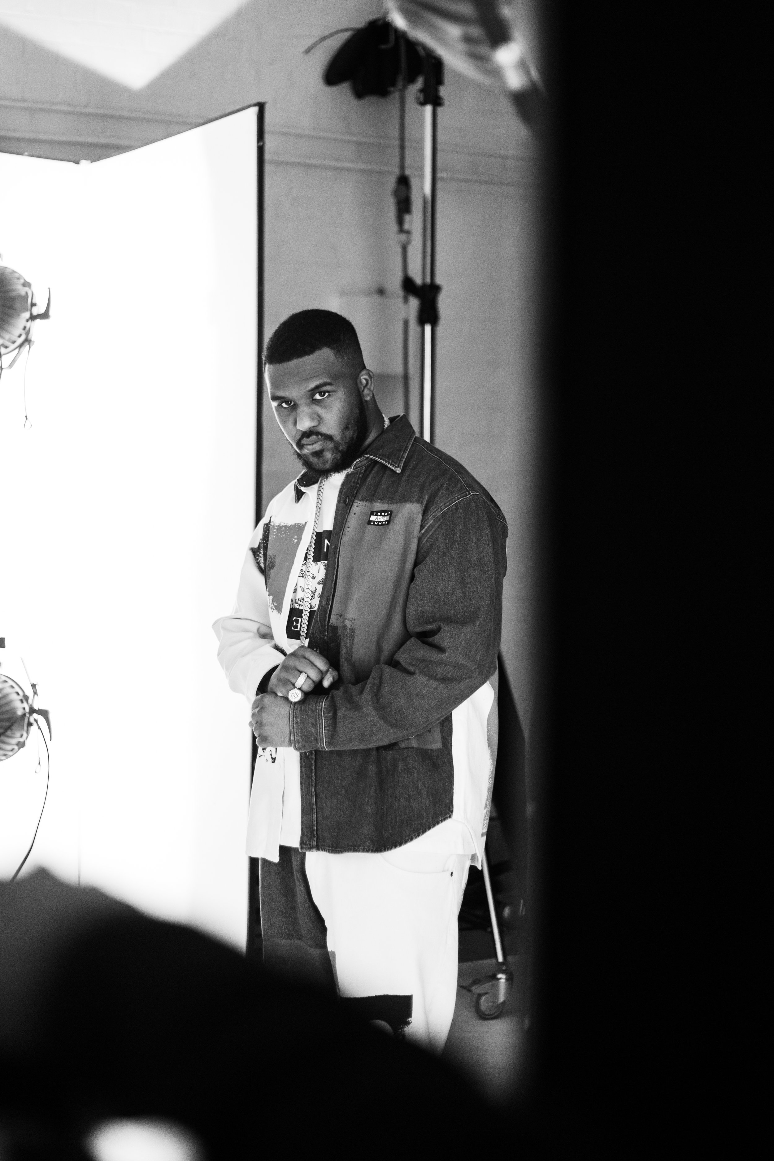 A man standing in a photography studio, wearing a denim jacket and posing for the camera, with photography equipment and a white backdrop visible in the background.