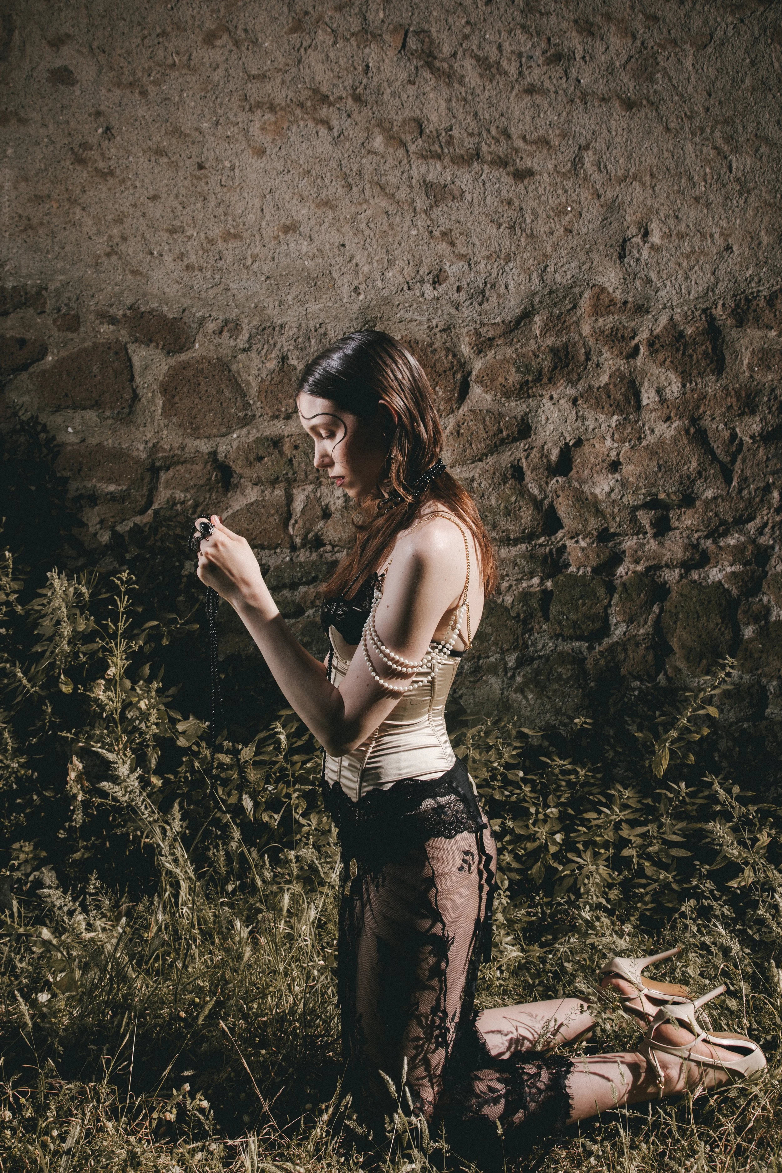 A woman dressed in black lace and a cream corset, kneeling on grass and holding a necklace, with a stone wall behind her and beige high heels on the ground nearby.