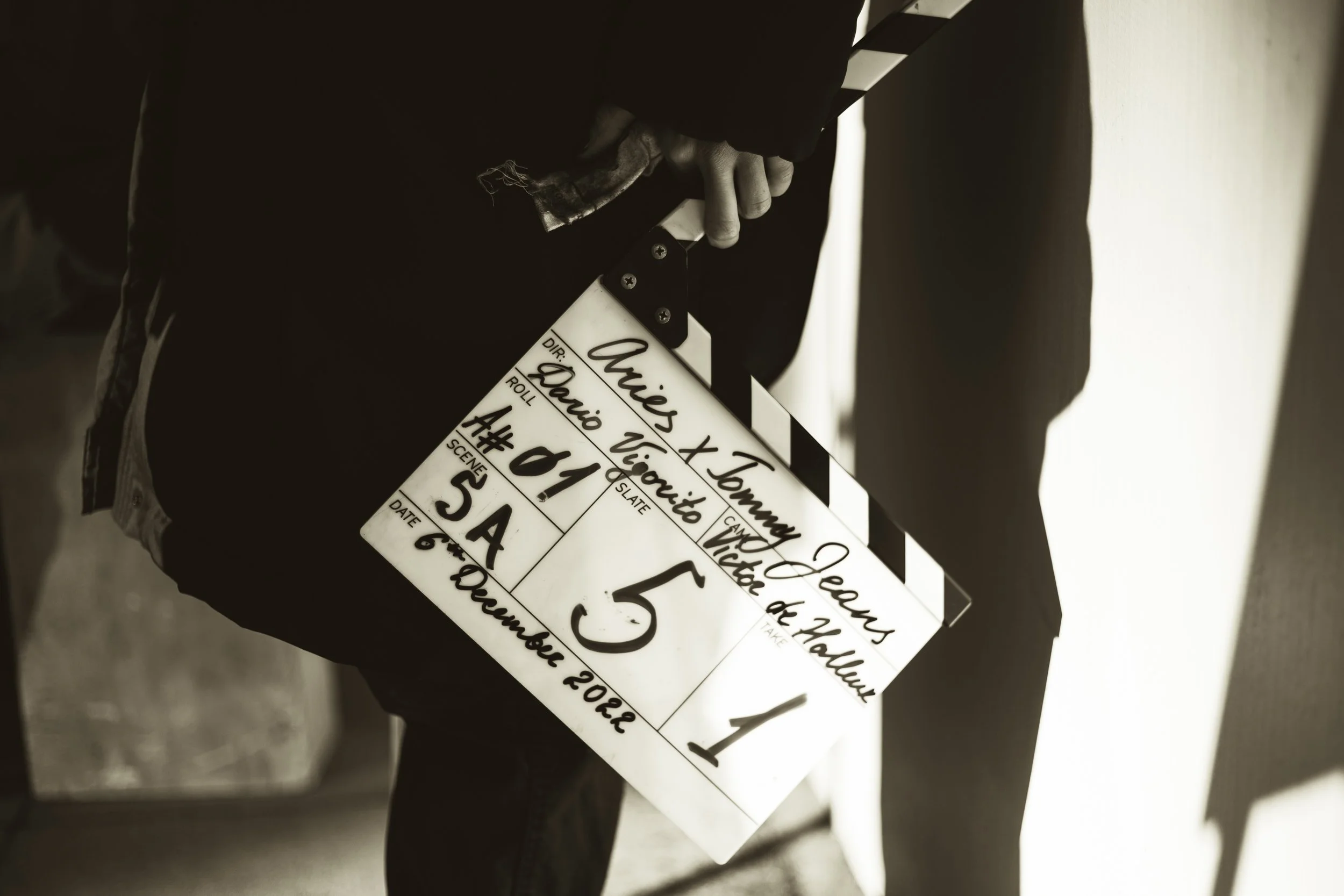 A person holding a director's clapperboard with handwritten details for a film scene, including names, scene number, and date.