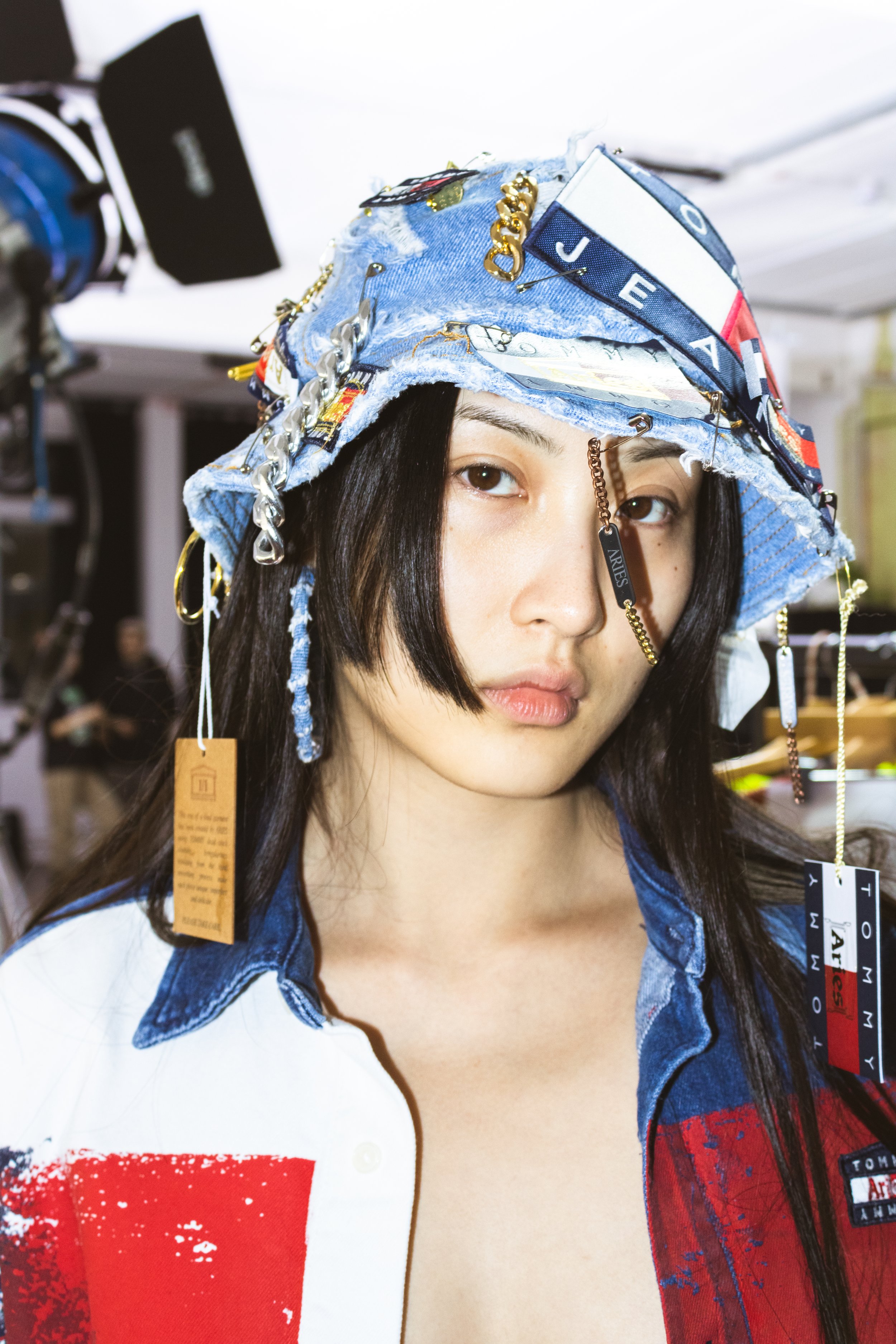 A young woman wearing a ripped denim bucket hat decorated with chains and tags, paired with a red, white, and blue fashion shirt, standing in a studio setting.