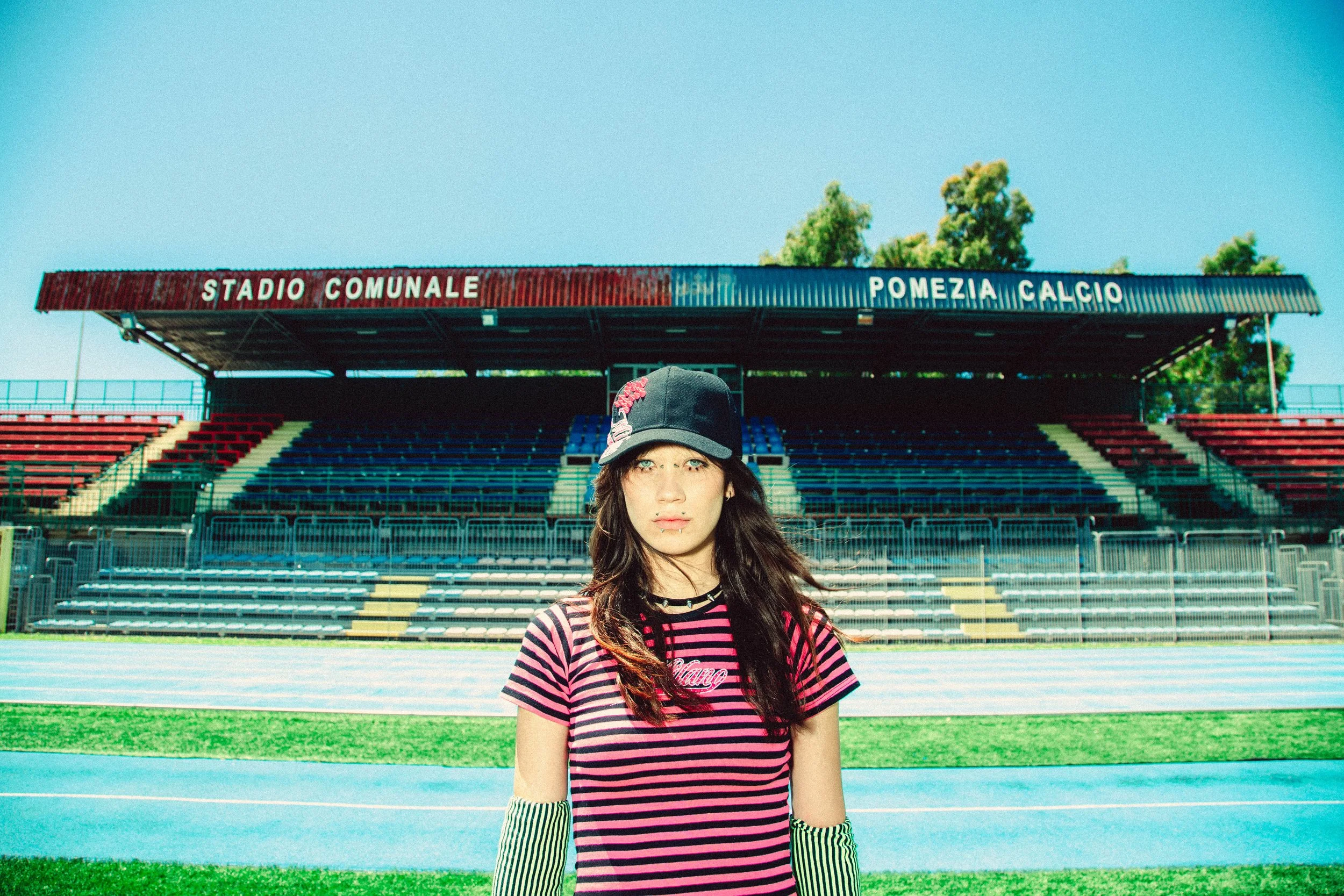 A young woman with long dark hair, wearing a striped pink and black t-shirt, striped arm warmers, a black cap, and a lip piercing, standing in front of an empty outdoor stadium with seating and a sign that reads "Stadio Comunale Pomezia Calcio" under