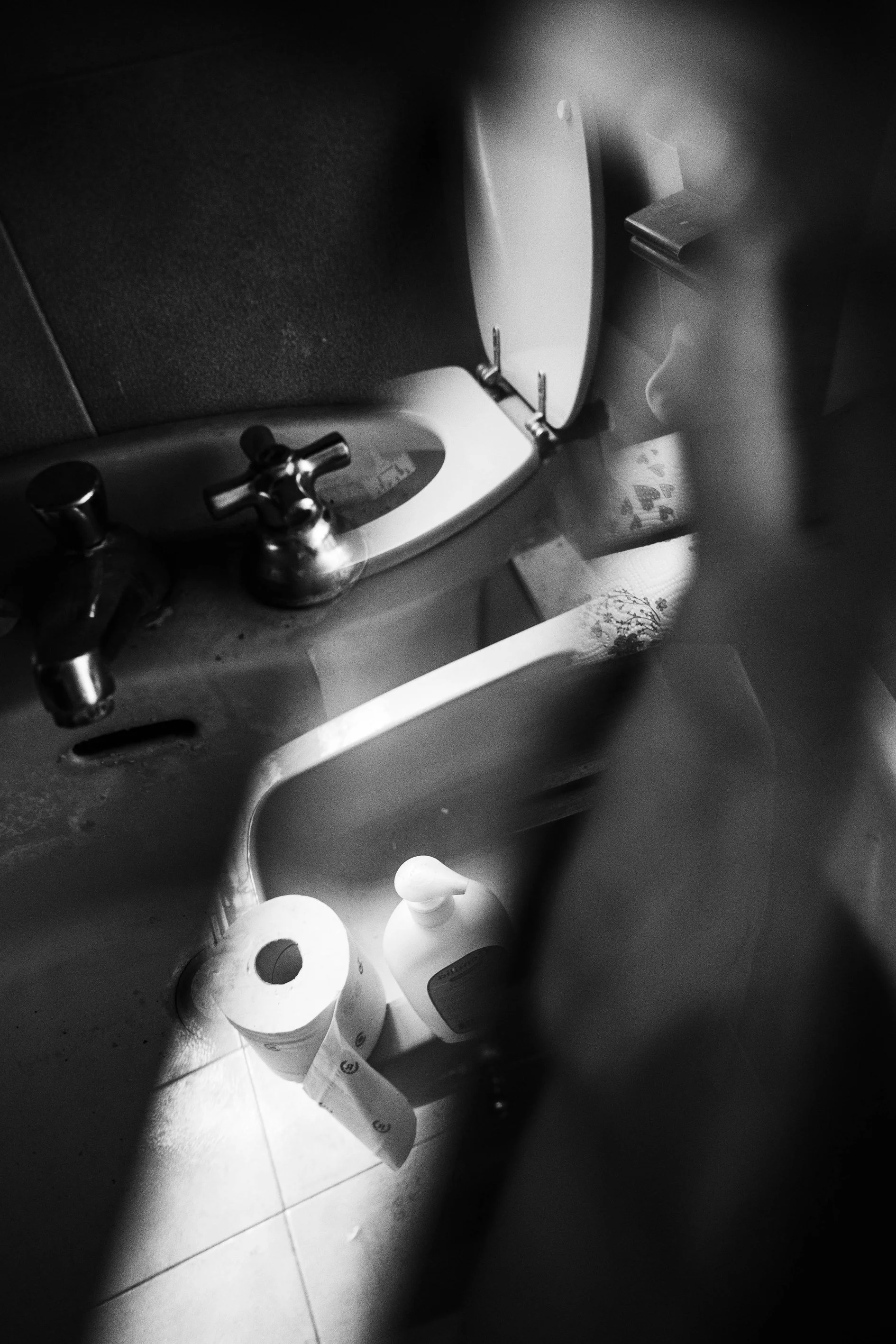 A black and white photo of a bathroom sink with an open mirror cabinet above, showing a reflection. The sink has two faucet handles, and a roll of toilet paper and a hand soap bottle are placed underneath it on the floor.