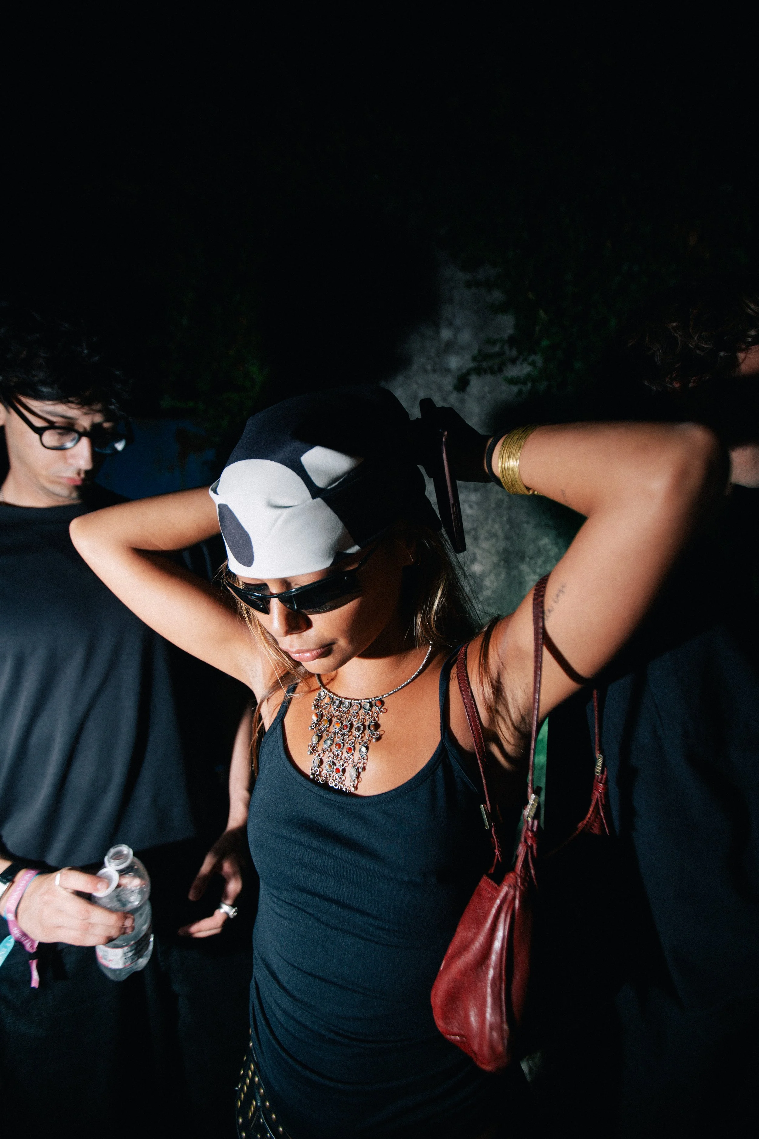 A woman wearing sunglasses, a black tank top, and a cow-print bandana over her hair, with jewelry and a red bag, is adjusting her hair with her hands behind her head. She is standing among people in a dark outdoor setting.