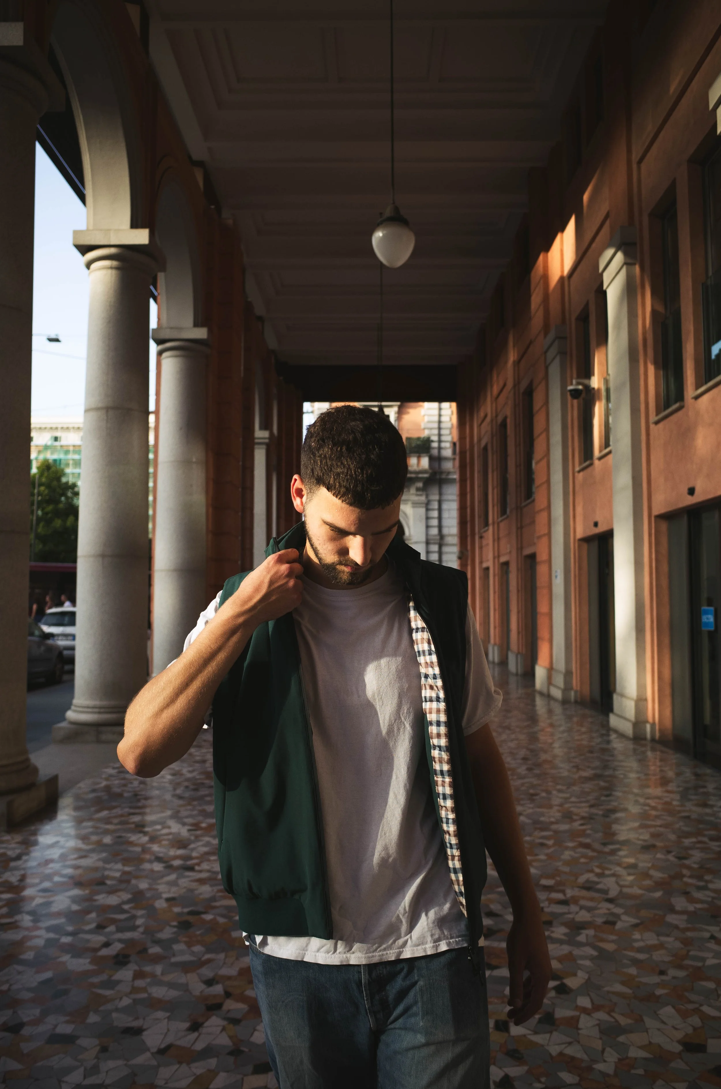 A young man walking outside in the city, adjusting his jacket over a white t-shirt, with a building and columns in the background.