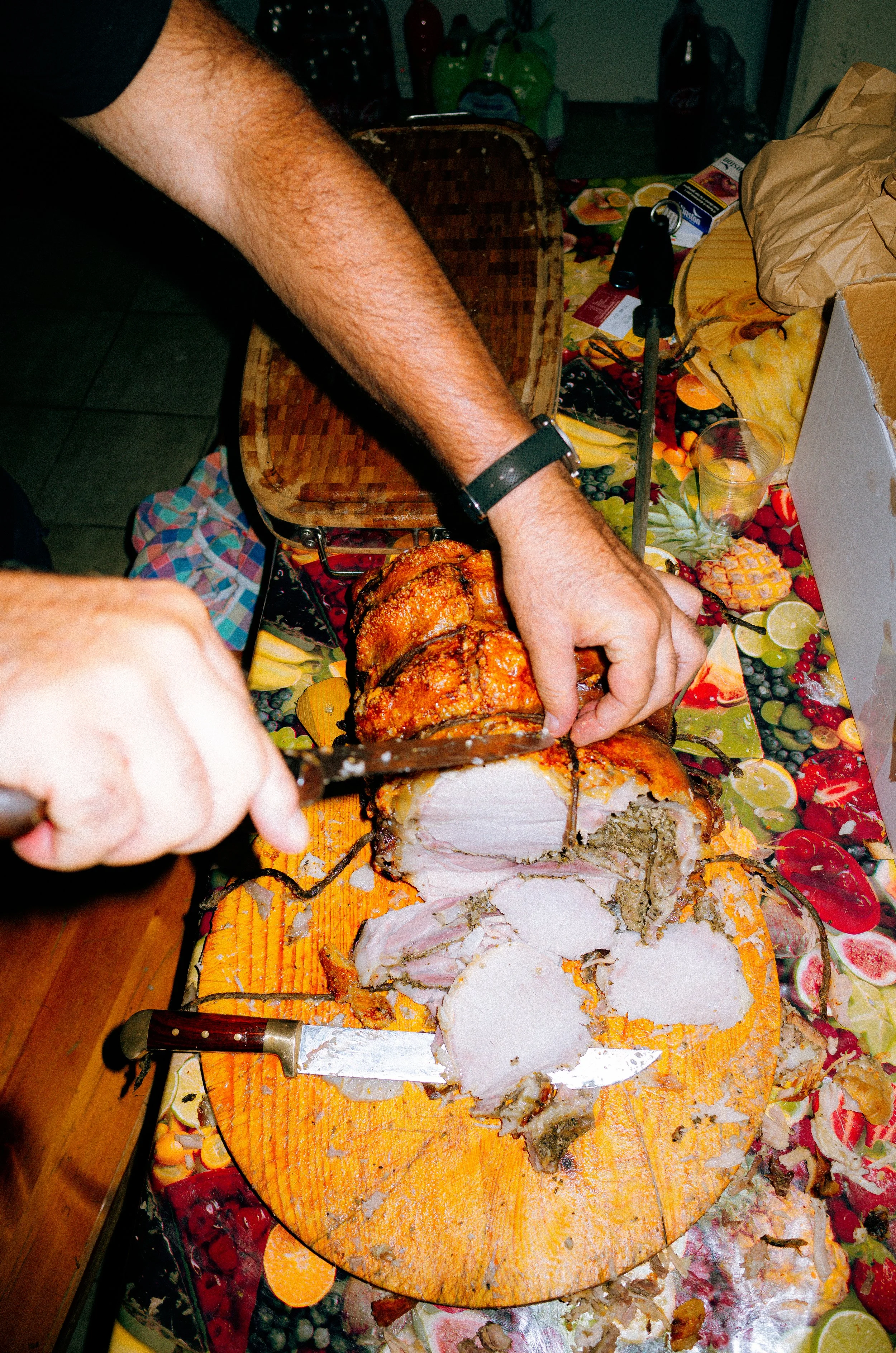 Person slicing a cooked meat roast on a wooden cutting board, with a tablecloth featuring fruit patterns in the background.